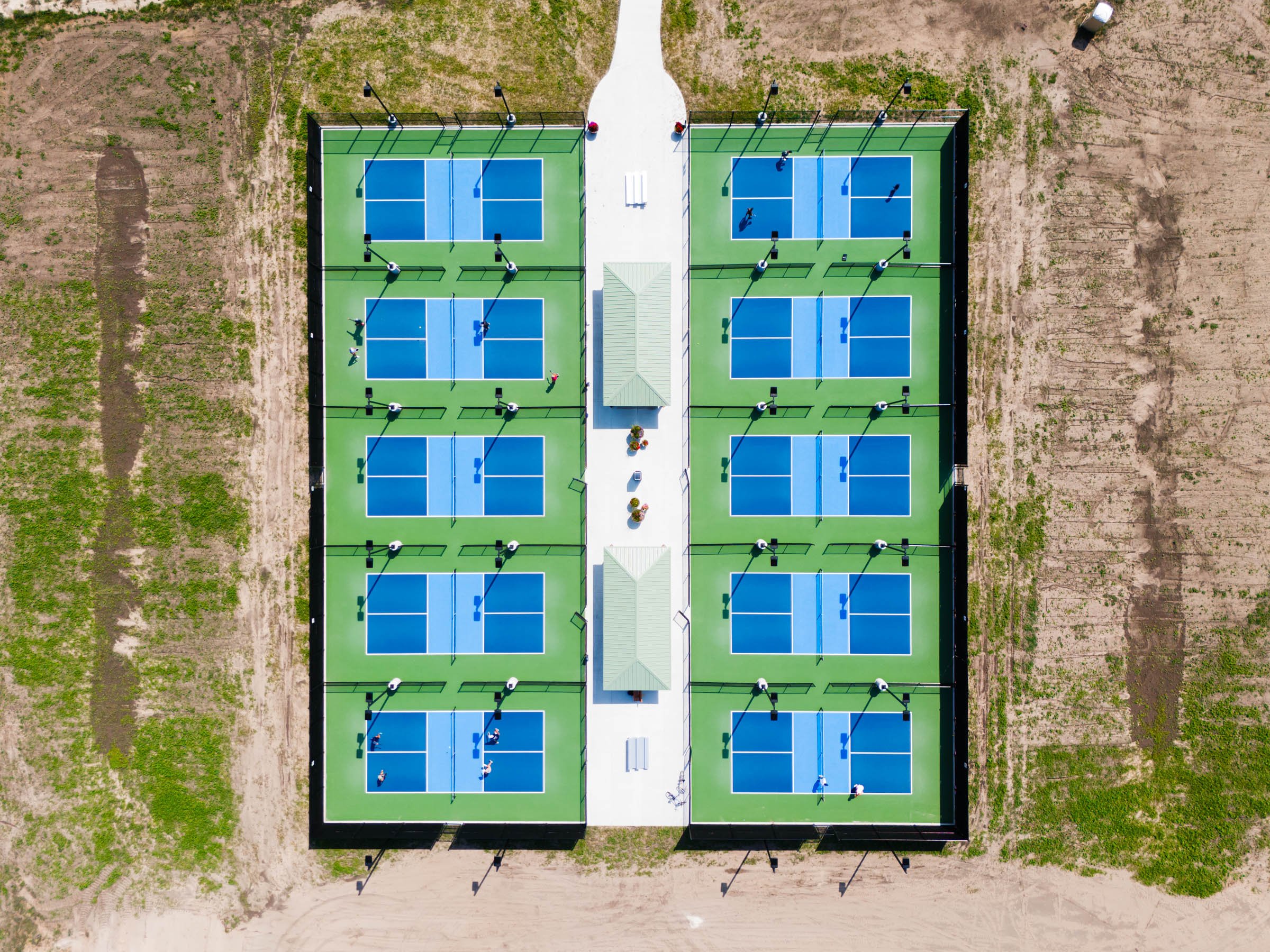 An aerial view of a ten pickleball court facility with two tone blue courts and green outside surface and fencing, with players on the courts and seating areas in the center.