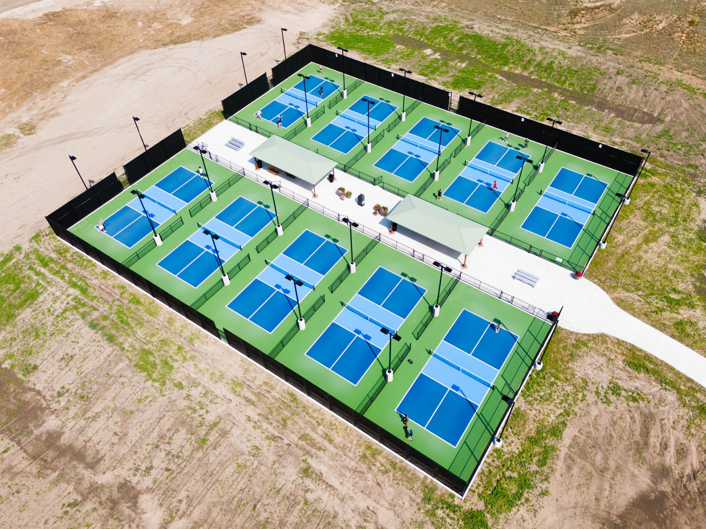 Aerial view of a fenced outdoor pickleball complex with multiple blue pickleball courts, surrounded by a dirt and grassy area, featuring pathways, benches, and two shaded pavilions.