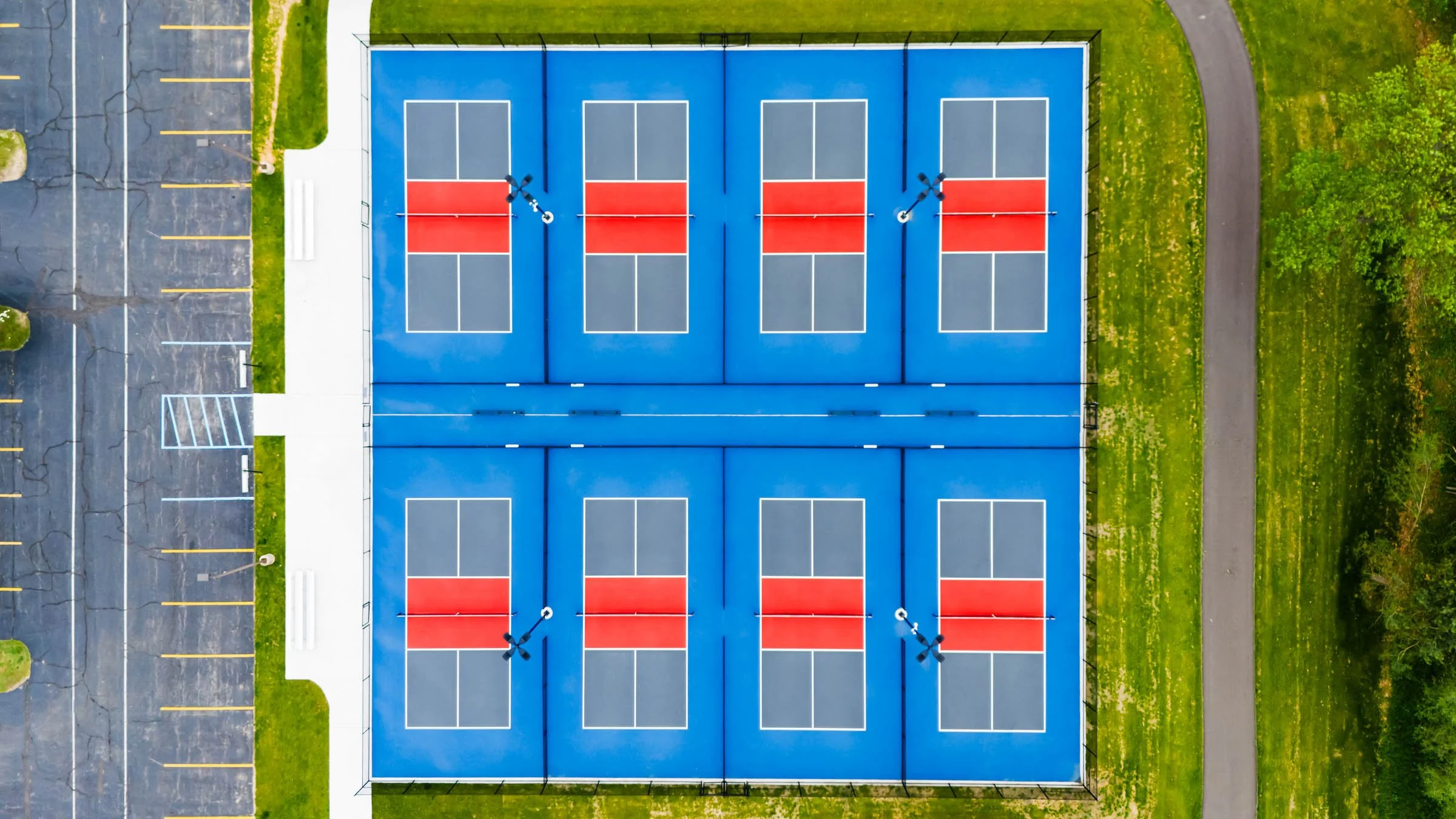 Aerial view of eight pickleball courts, in Cannon Township, with blue surface and red playing areas, surrounded by green grass and a parking lot.