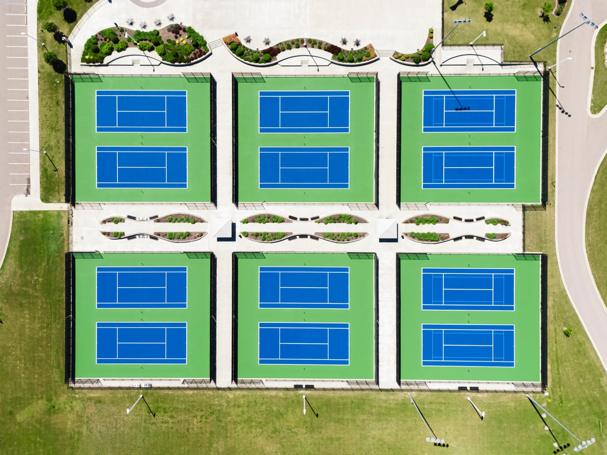 Aerial view of twelve tennis courts in, Byron Center Michigan, arranged in two rows with a landscaped walkway in the middle, surrounded by a parking lot and grassy areas.