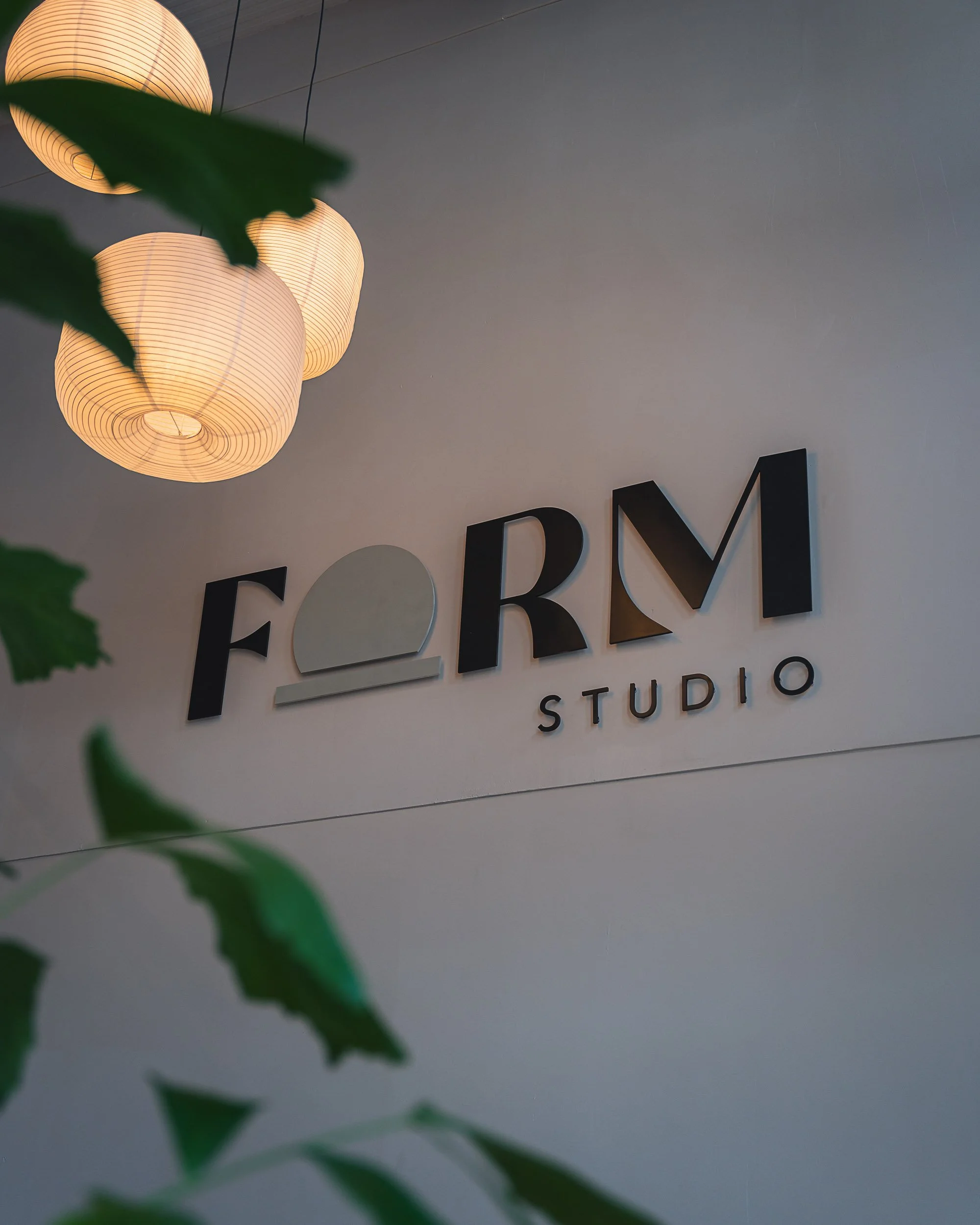 Interior view of FORM Studio sign on wall, illuminated paper lanterns overhead, with green leaves partially visible in foreground.