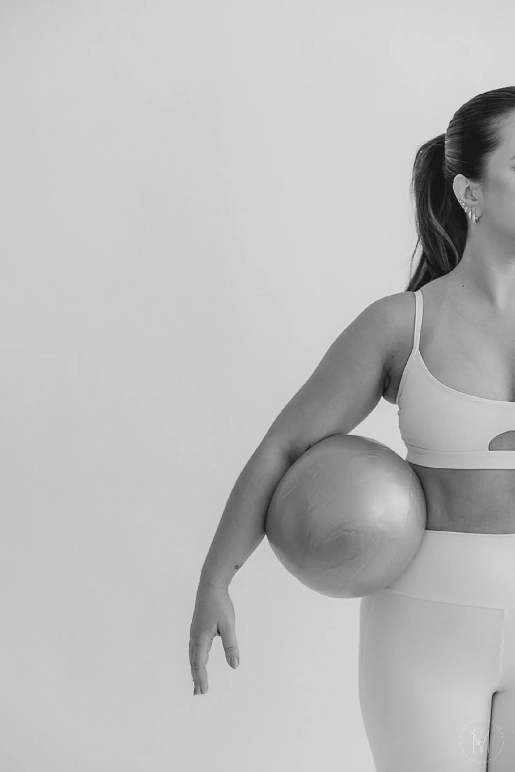 A woman with a ponytail wearing fitness attire holding a large exercise ball against her side in a minimalist setting.