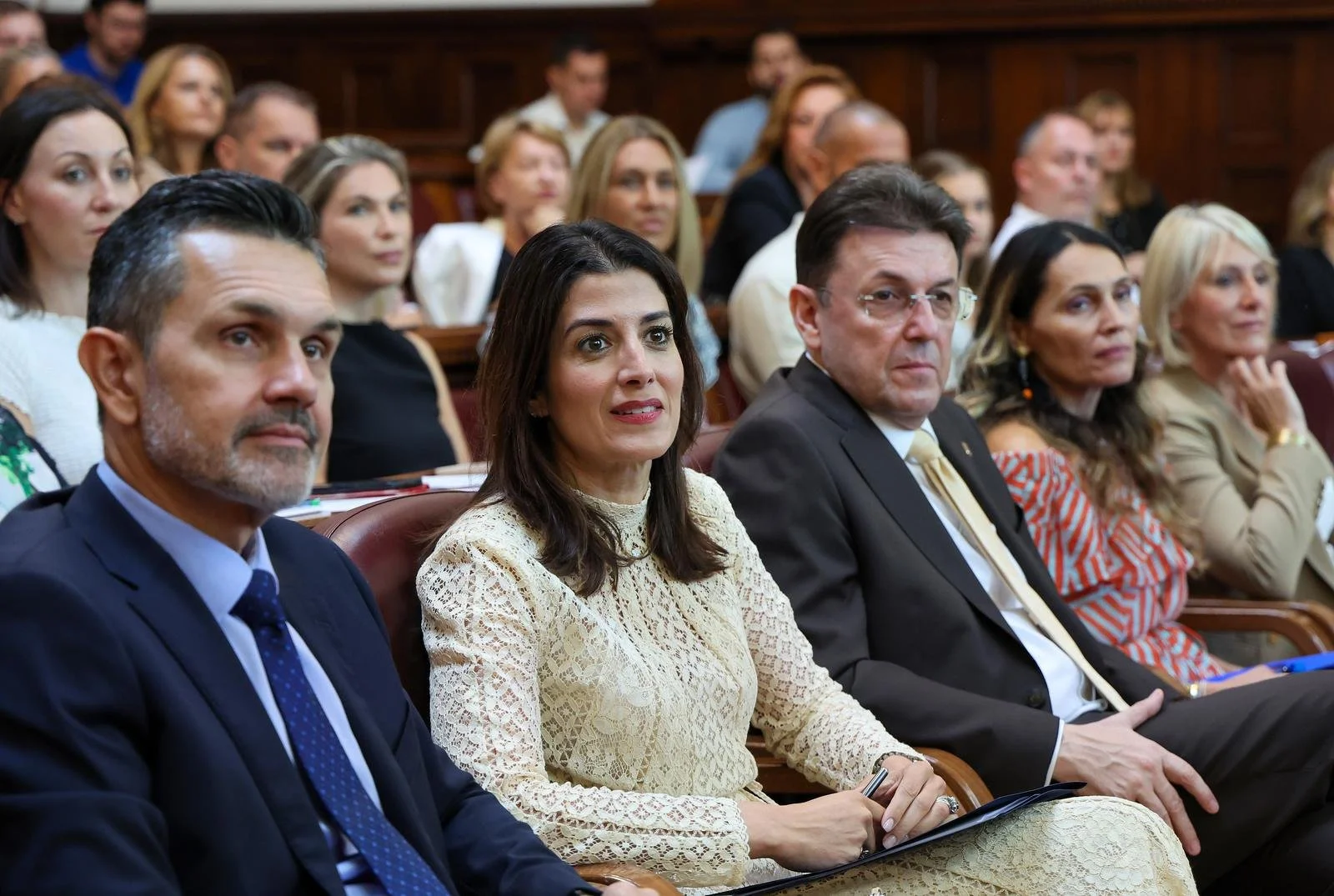Ambassador Nathalie Rayes attending a formal event, seated in rows, wearing business attire, focused on a presentation.