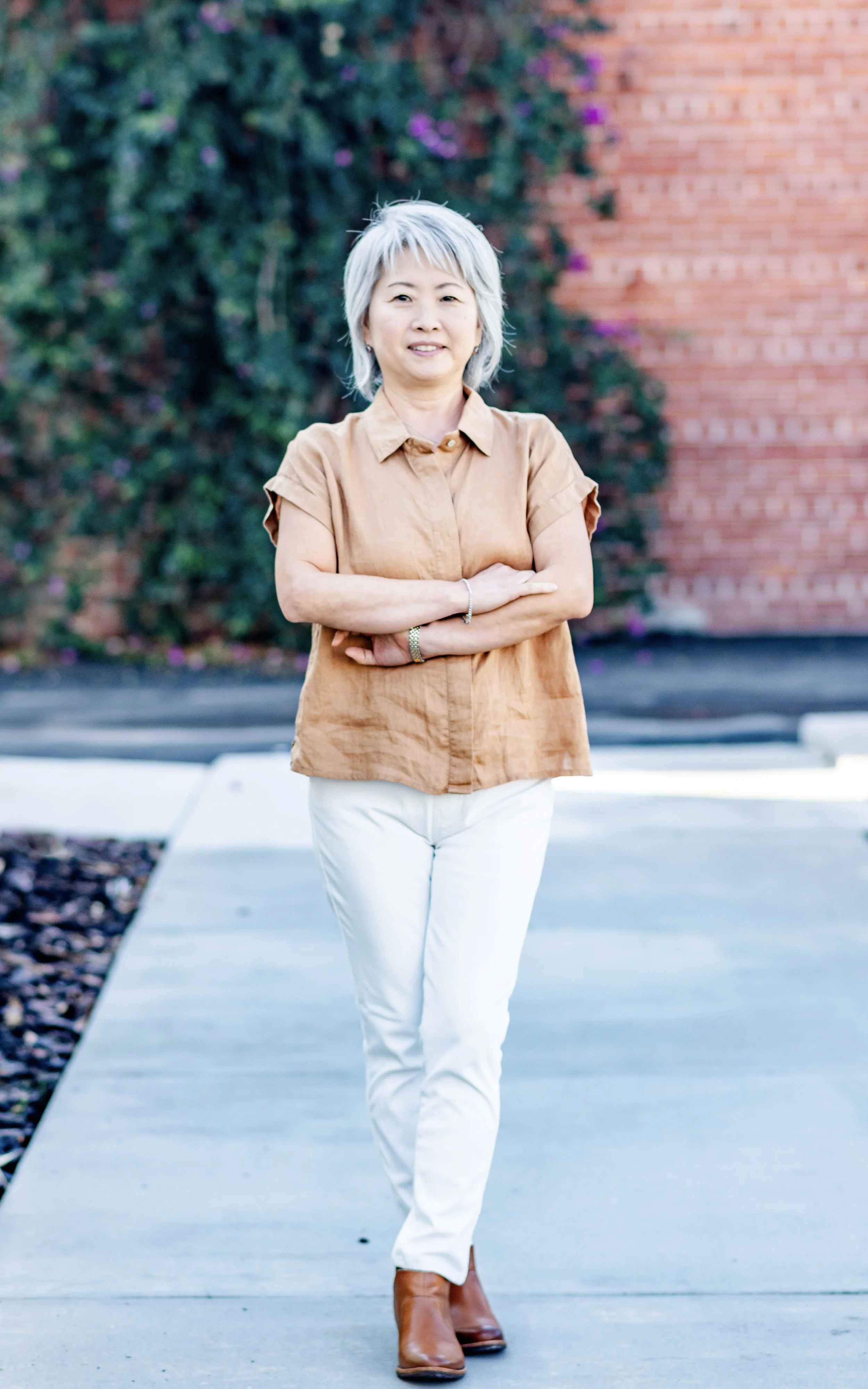 A middle-aged woman with gray hair standing outdoors with her arms crossed, wearing a brown short-sleeve shirt, white pants, and brown ankle boots, with a background of a brick wall and greenery.