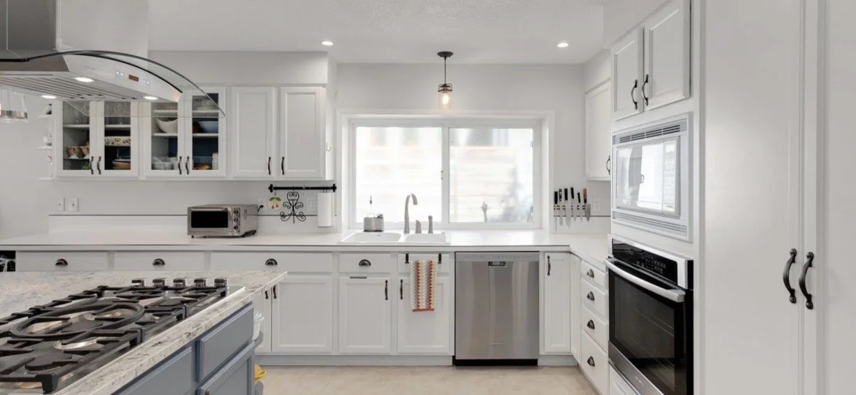 Clean, white kitchen with stainless steel appliances, a gas stove, microwave, dishwasher, and an overhead microwave oven, with a window above the sink and various kitchen utensils and decor.