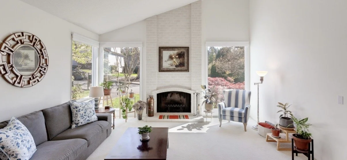 Bright living room with a fireplace, large windows, a gray sofa with cushions, a striped armchair, potted plants, and decorative items.