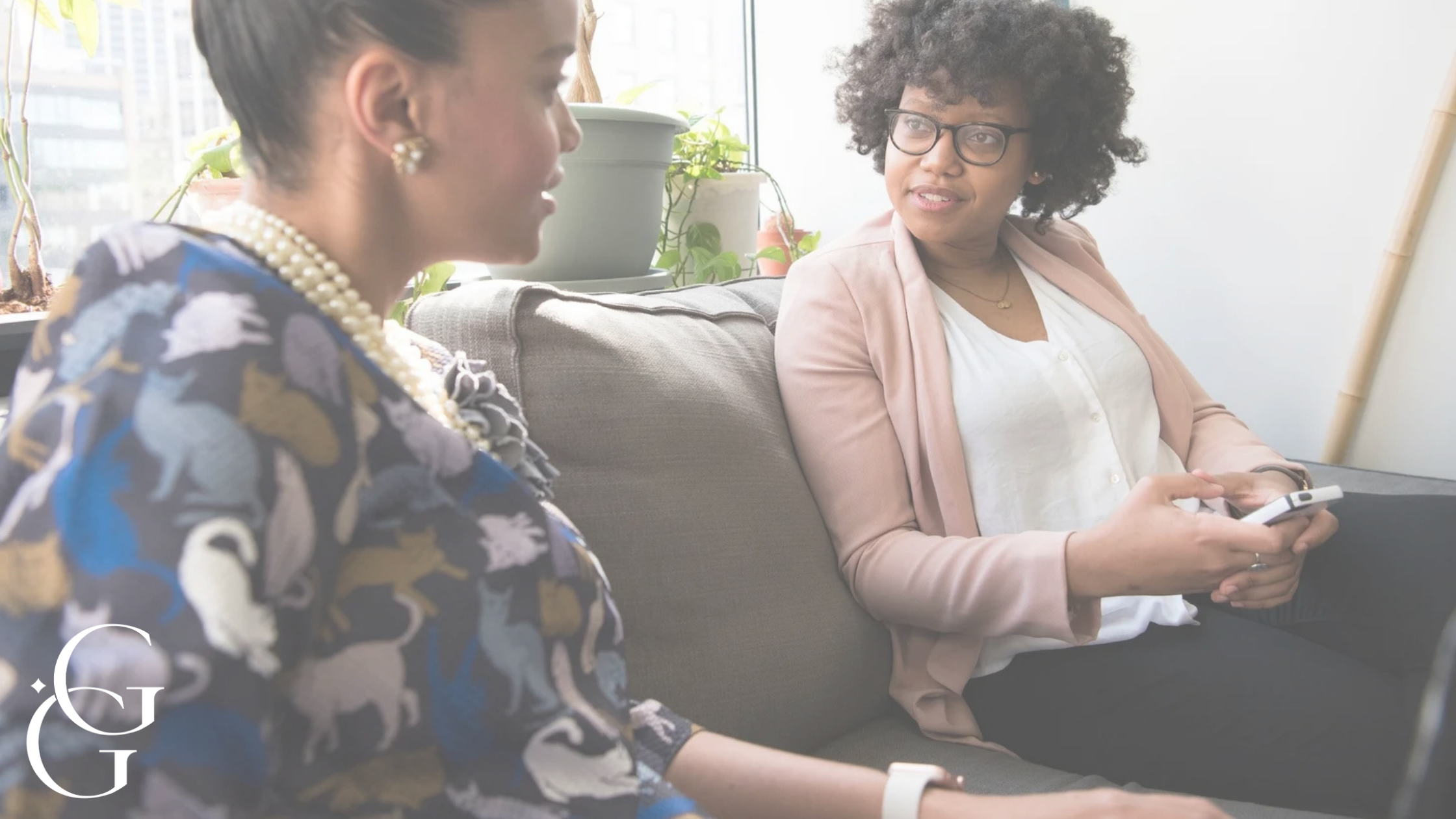 Two women sitting on a couch in an office, engaged in conversation. One woman has dark hair styled in an updo, wearing a patterned blazer and pearl jewelry. The other woman has curly hair, glasses, and is holding a smartphone, wearing a light pink blazer and white top. Office plants and windows are in the background.