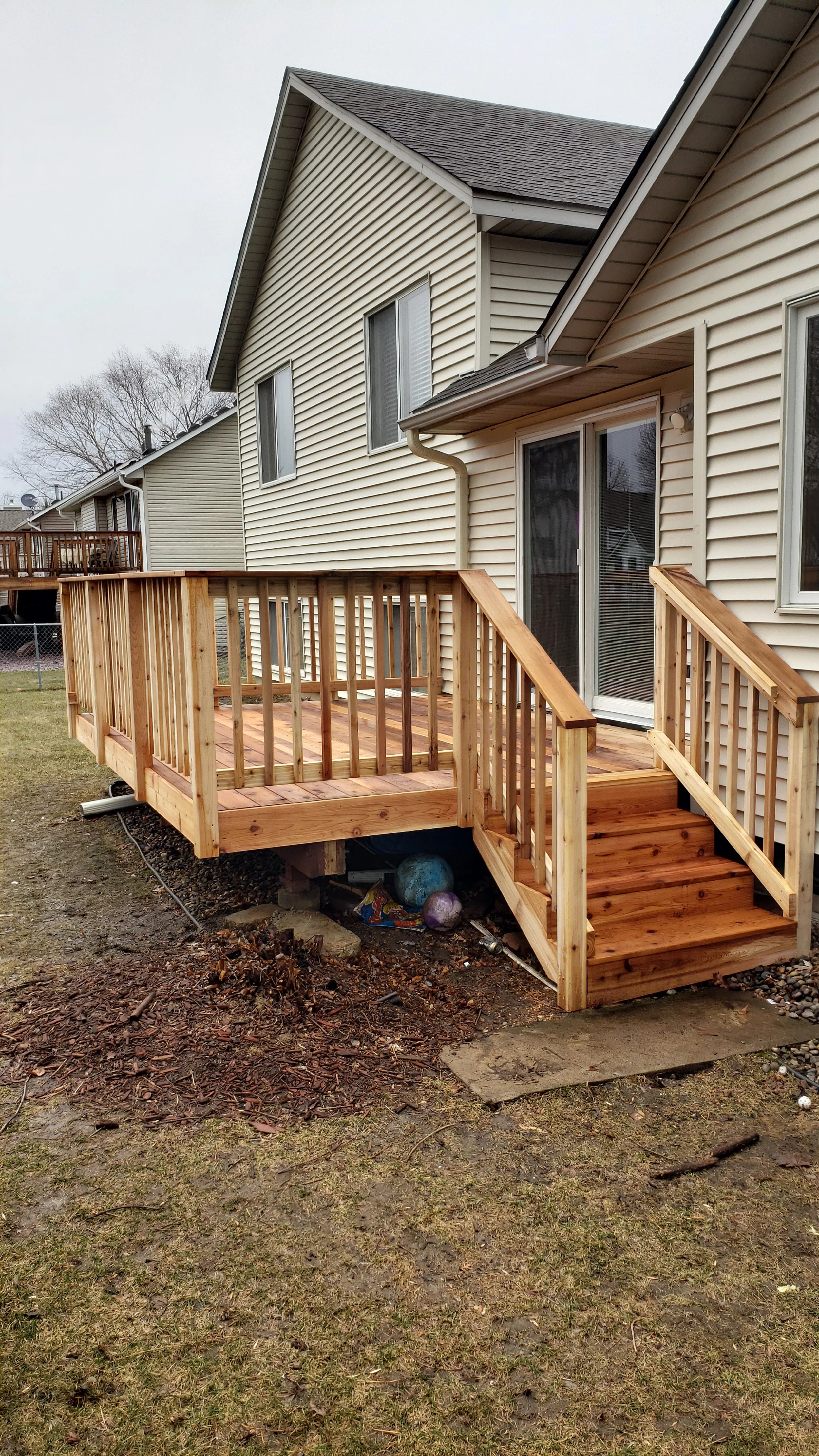 A newly built wooden deck attached to a beige house with sliding glass doors, surrounded by a grassy yard and other houses in the background.