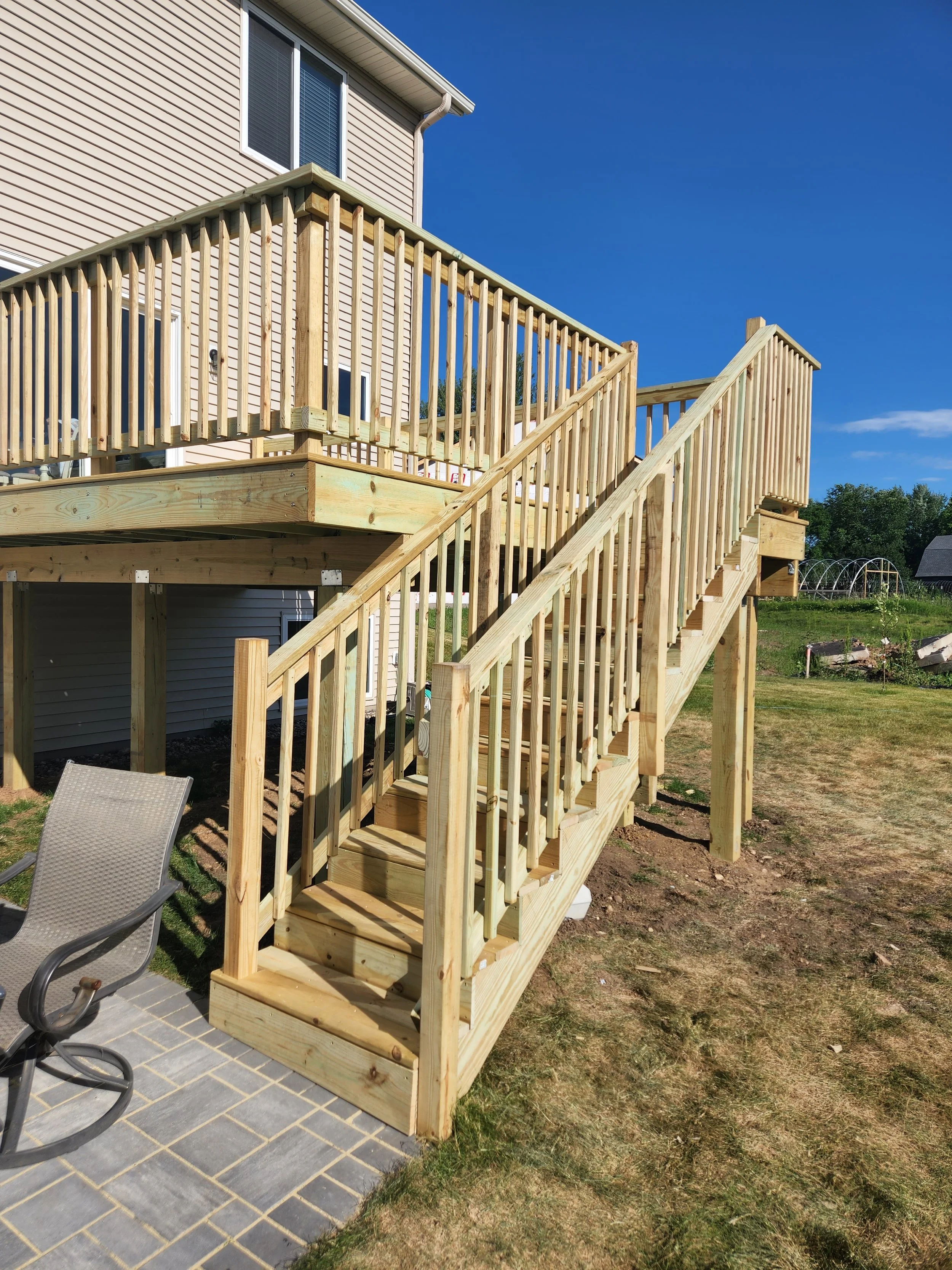 Newly built wooden staircase and deck attached to a beige house with beige siding, leading from a brick patio to the backyard, with a blue sky in the background.