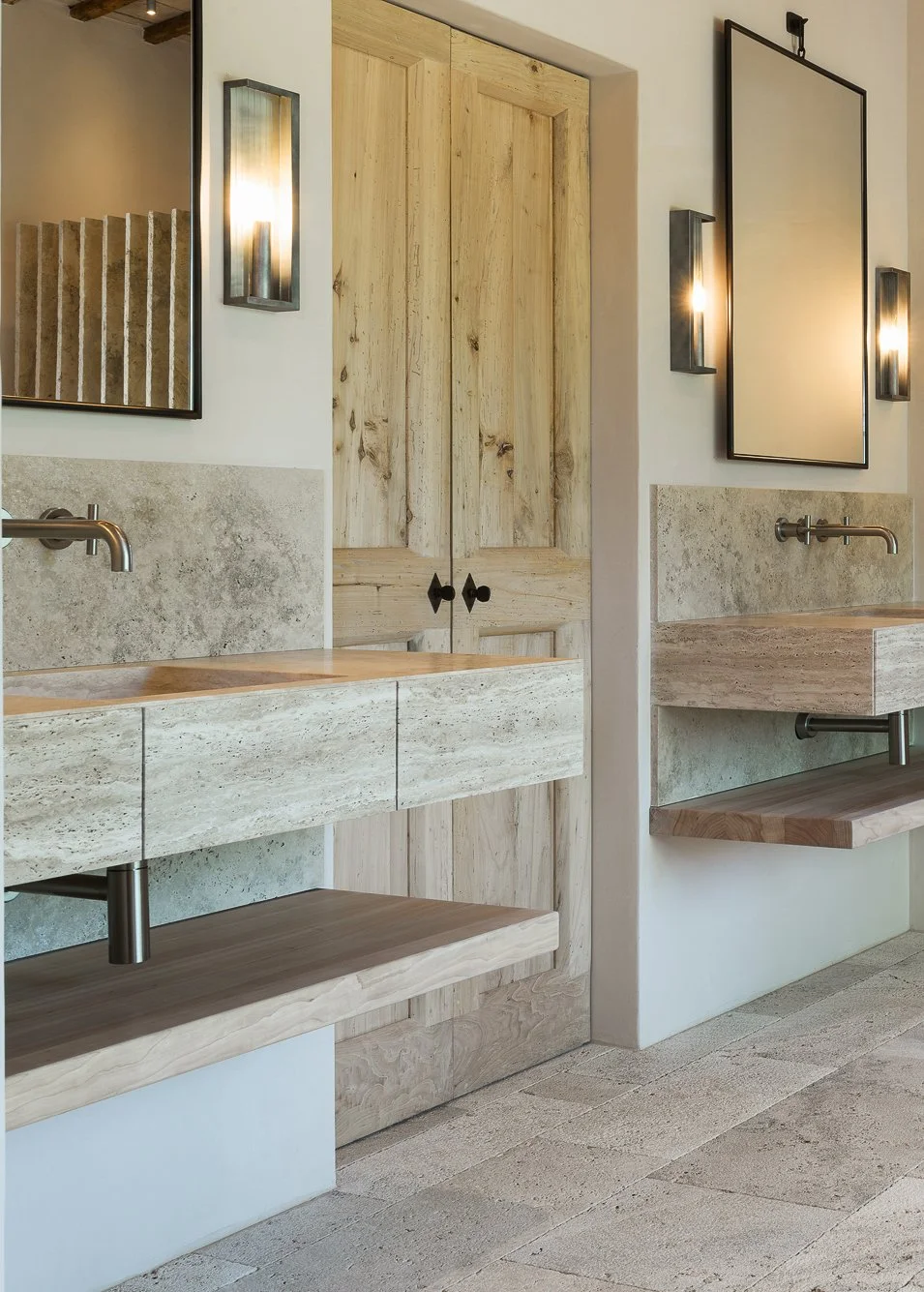 Two modern bathroom sinks with stone countertops, wall-mounted faucets, mirrors framed in black, wooden cabinets, and wall-mounted light fixtures, with beige tiled flooring and a wooden door in the background.