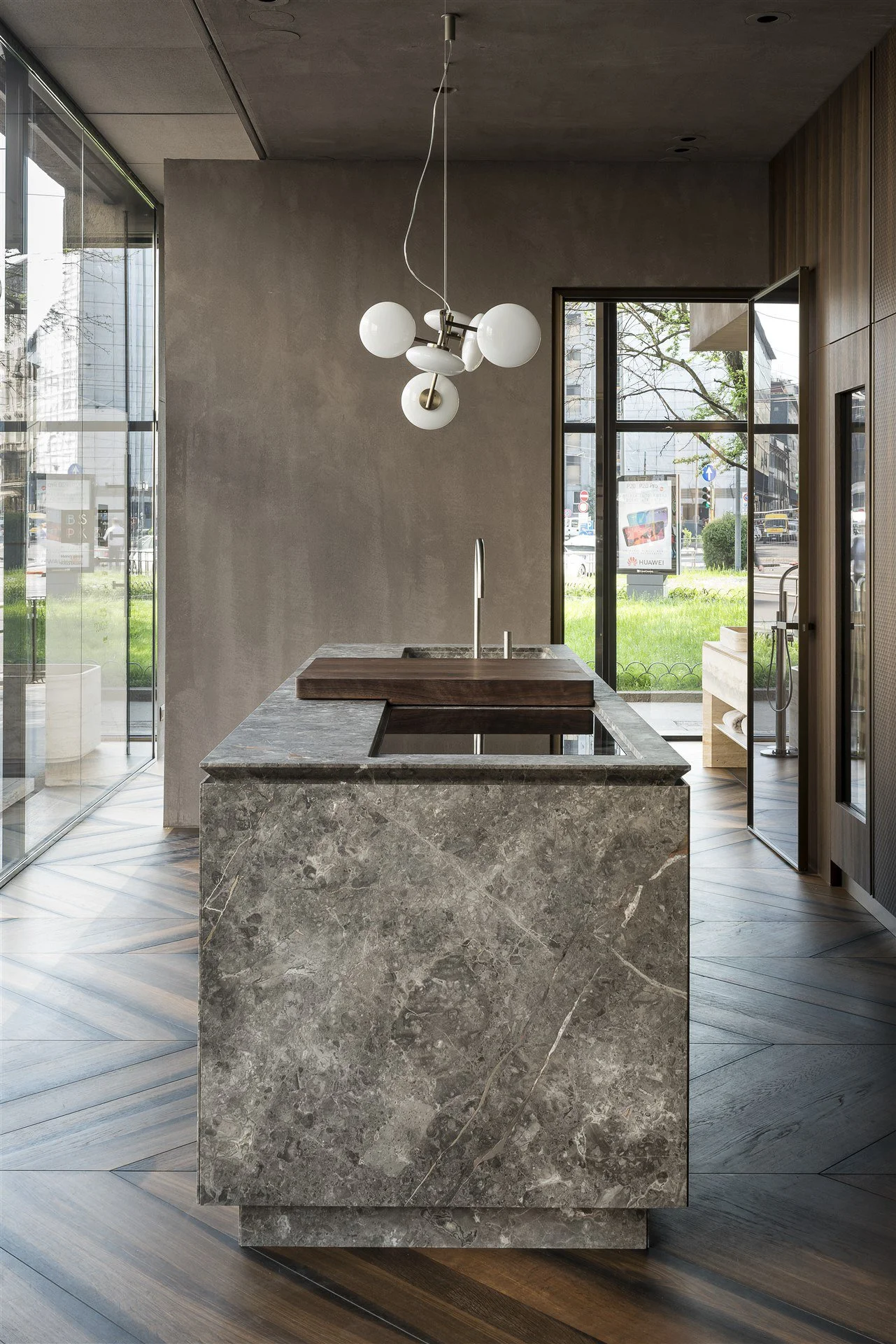 Modern kitchen island with gray marble finish, wooden cutting board, and a sink with faucet inside a contemporary space with large glass windows and wooden walls.
