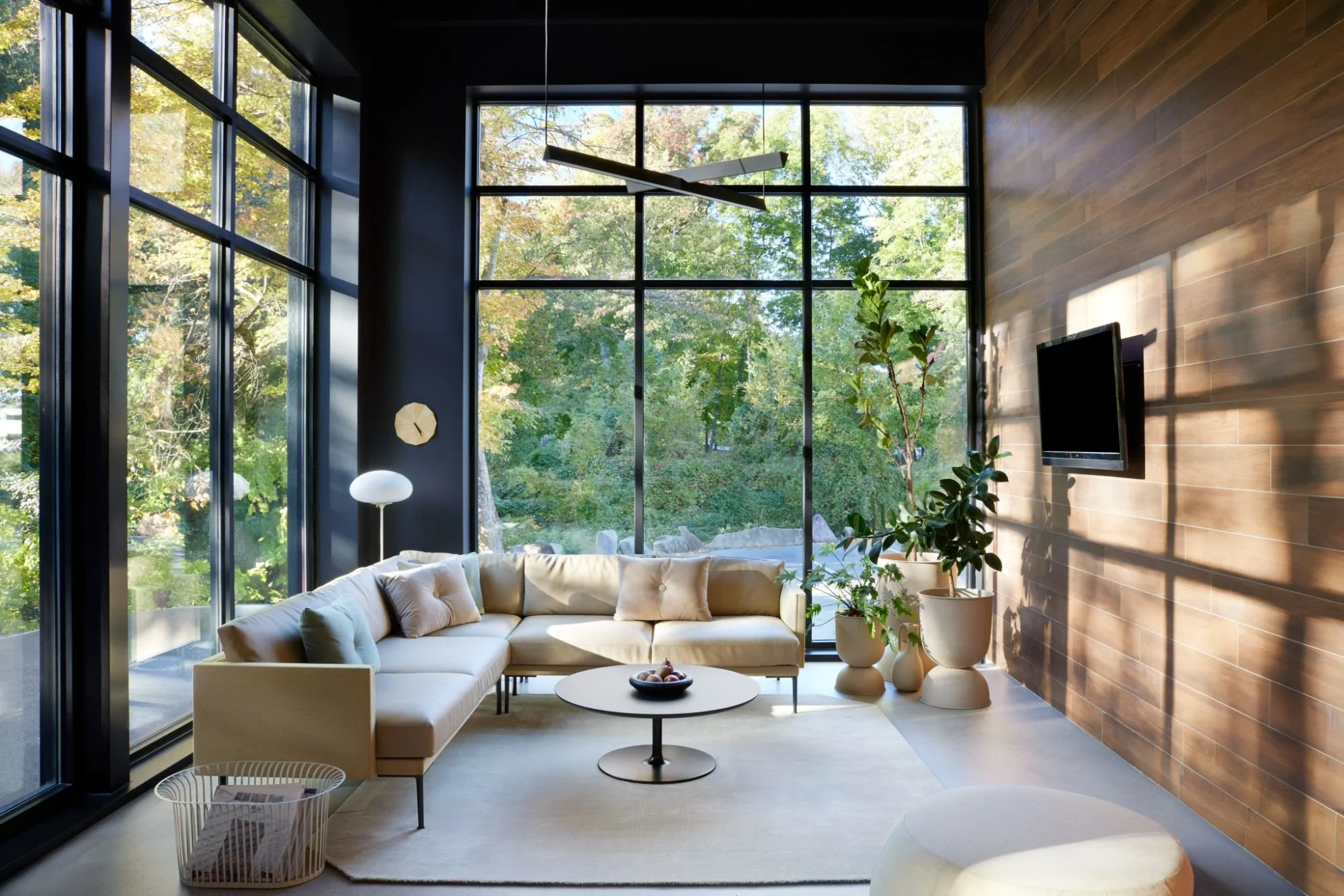 Sunlit commercial lounge with floor-to-ceiling windows, neutral sectional, modern decor, and lush greenery.