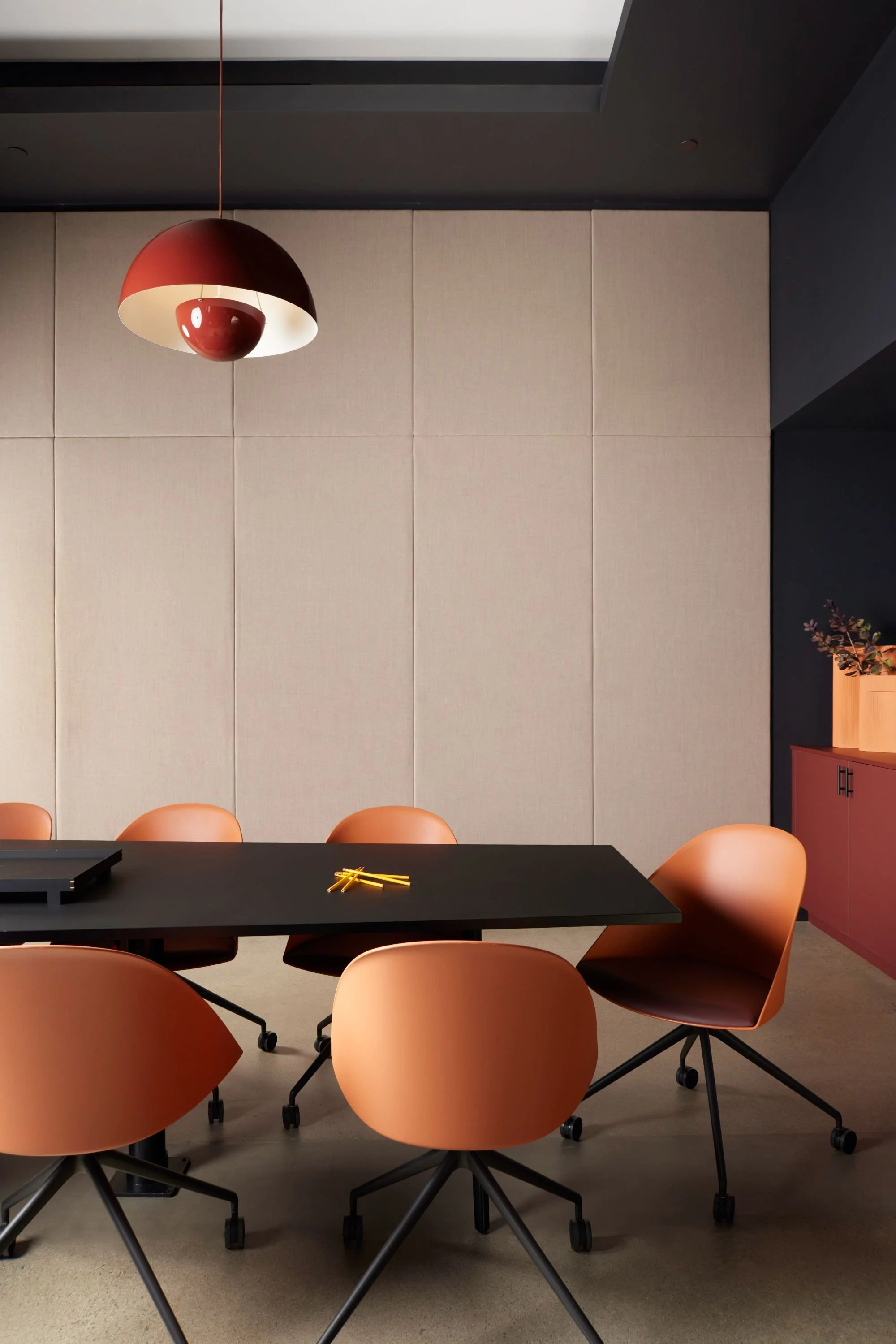 Modern meeting room with a black table, burnt-orange swivel chairs, custom upholstered walls, and a red pendant light.