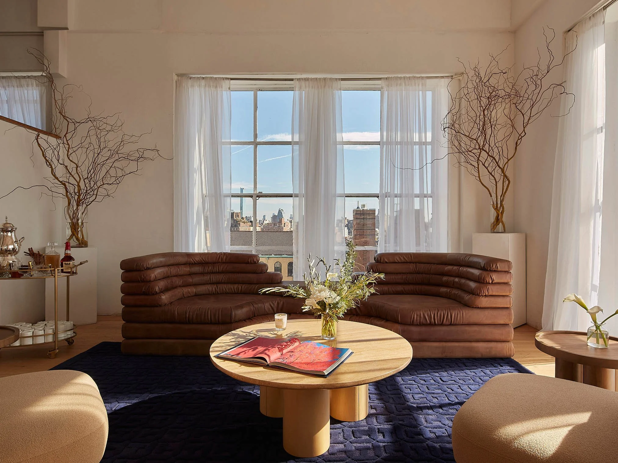 Luxury showroom with brown leather sofas, blue vintage rug, sculptural decor, and panoramic city views through large windows.