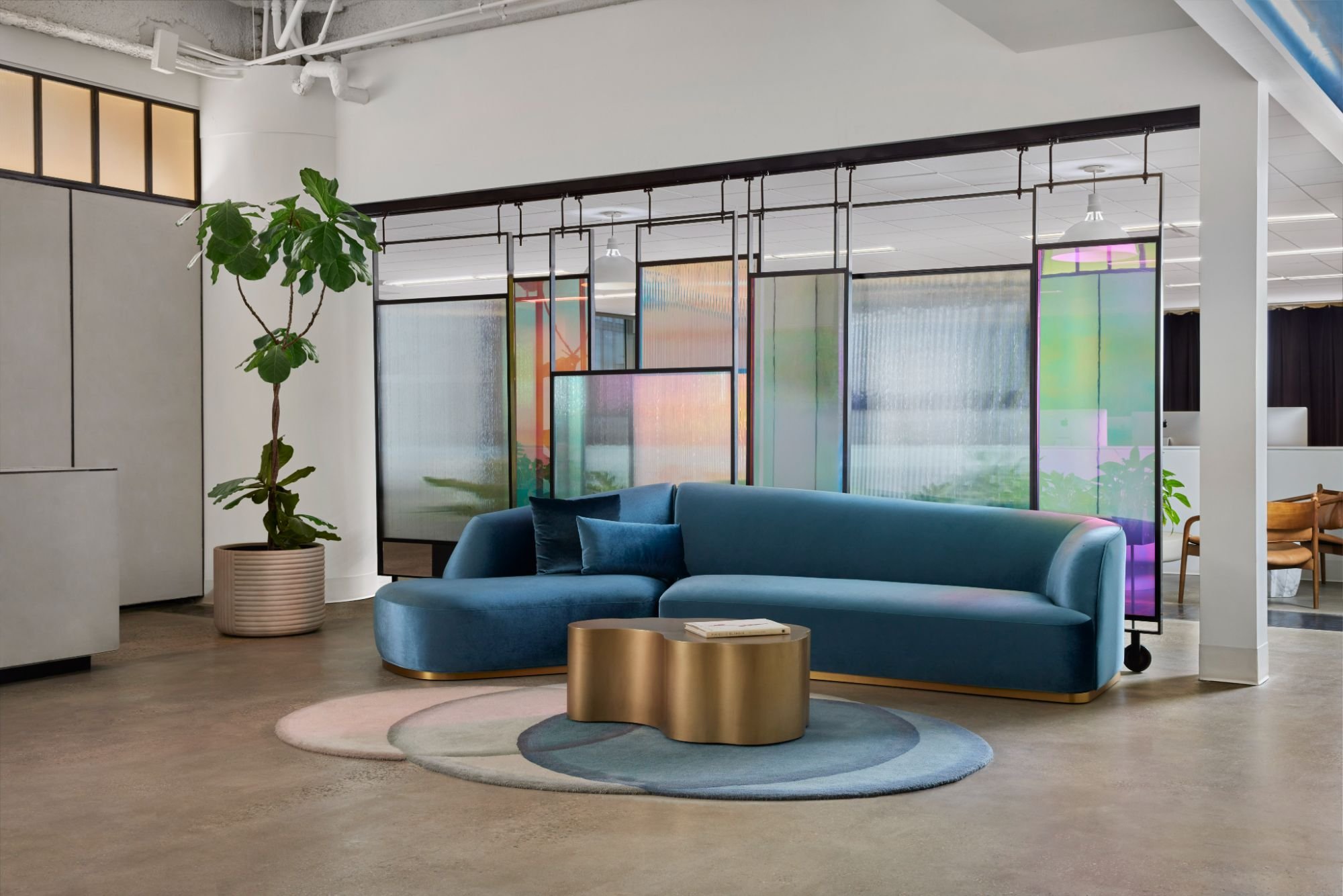 Modern office lobby with a curved blue velvet sofa, gold modern coffee table, and iridescent glass partitions styled with a fig tree.