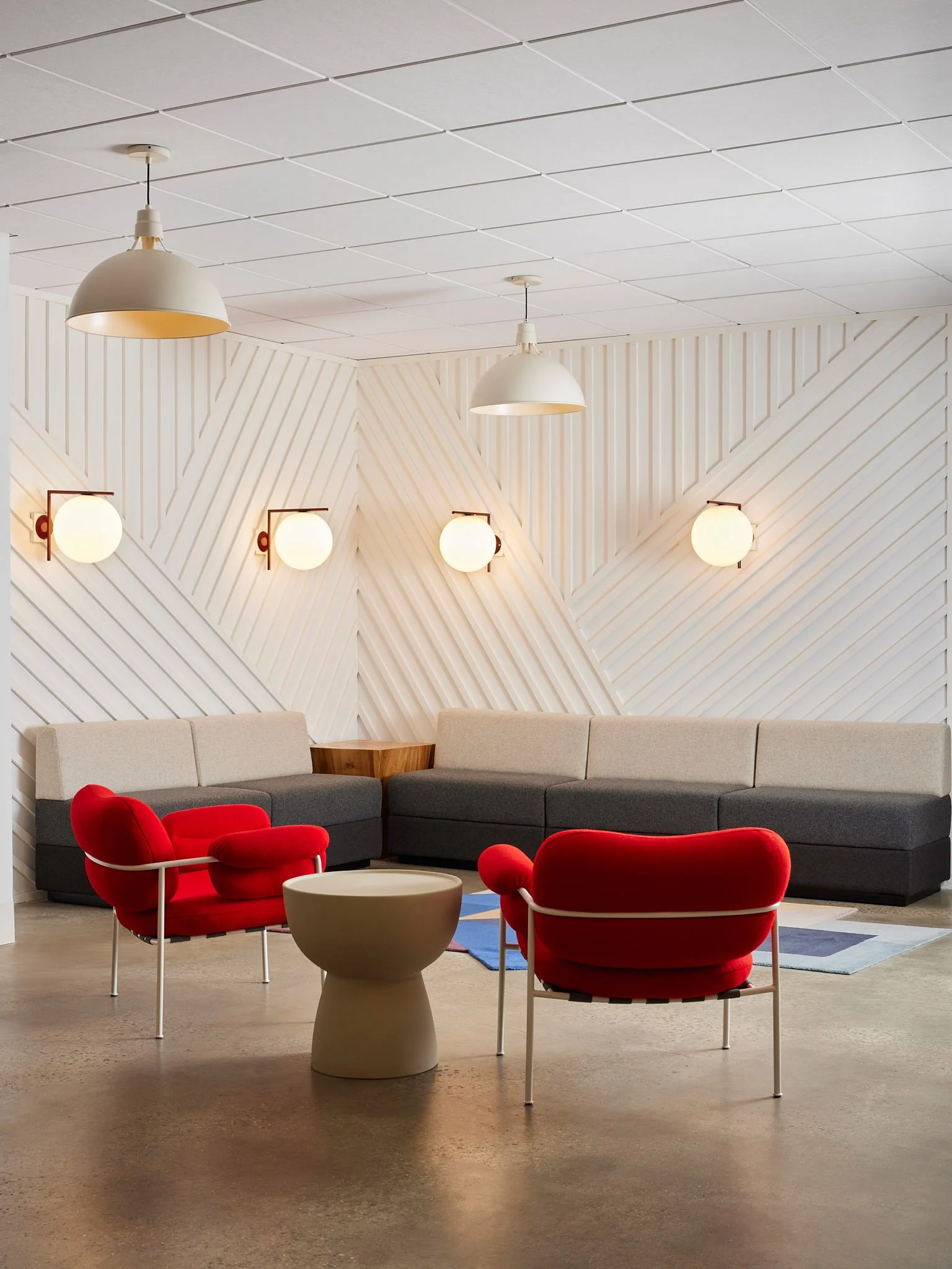 Modern office lounge with red accent chairs, a neutral sectional, sculptural lighting, and geometric wood wall panels.