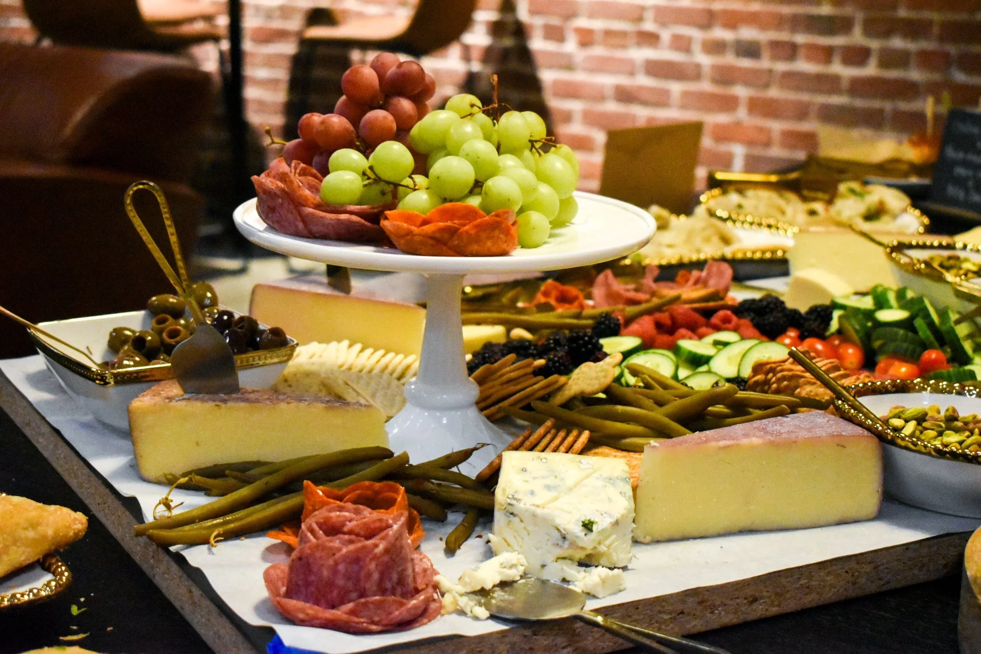 A cheese and fruit platter with grapes, olives, sliced vegetables, and various cheeses on a wooden board.