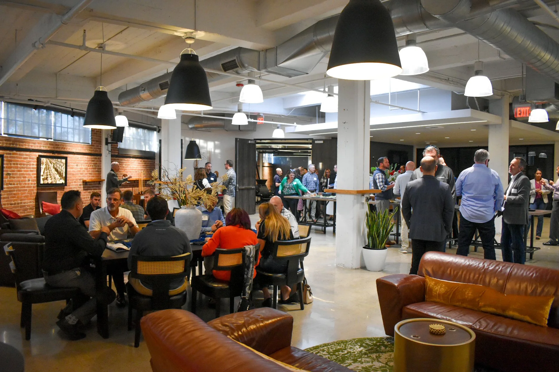 Indoor gathering at a modern event space or restaurant, with people sitting at tables and standing, brick walls, large hanging black and white pendant lights, and potted plants.