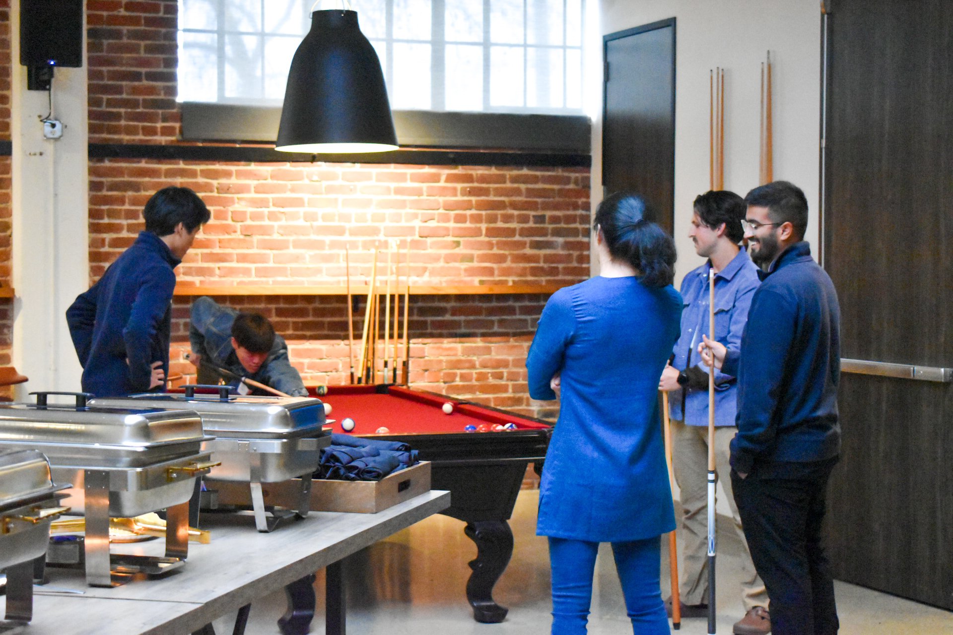 Group of five young adults watching a pool game in a modern, industrial-style room with brick wall and large window.