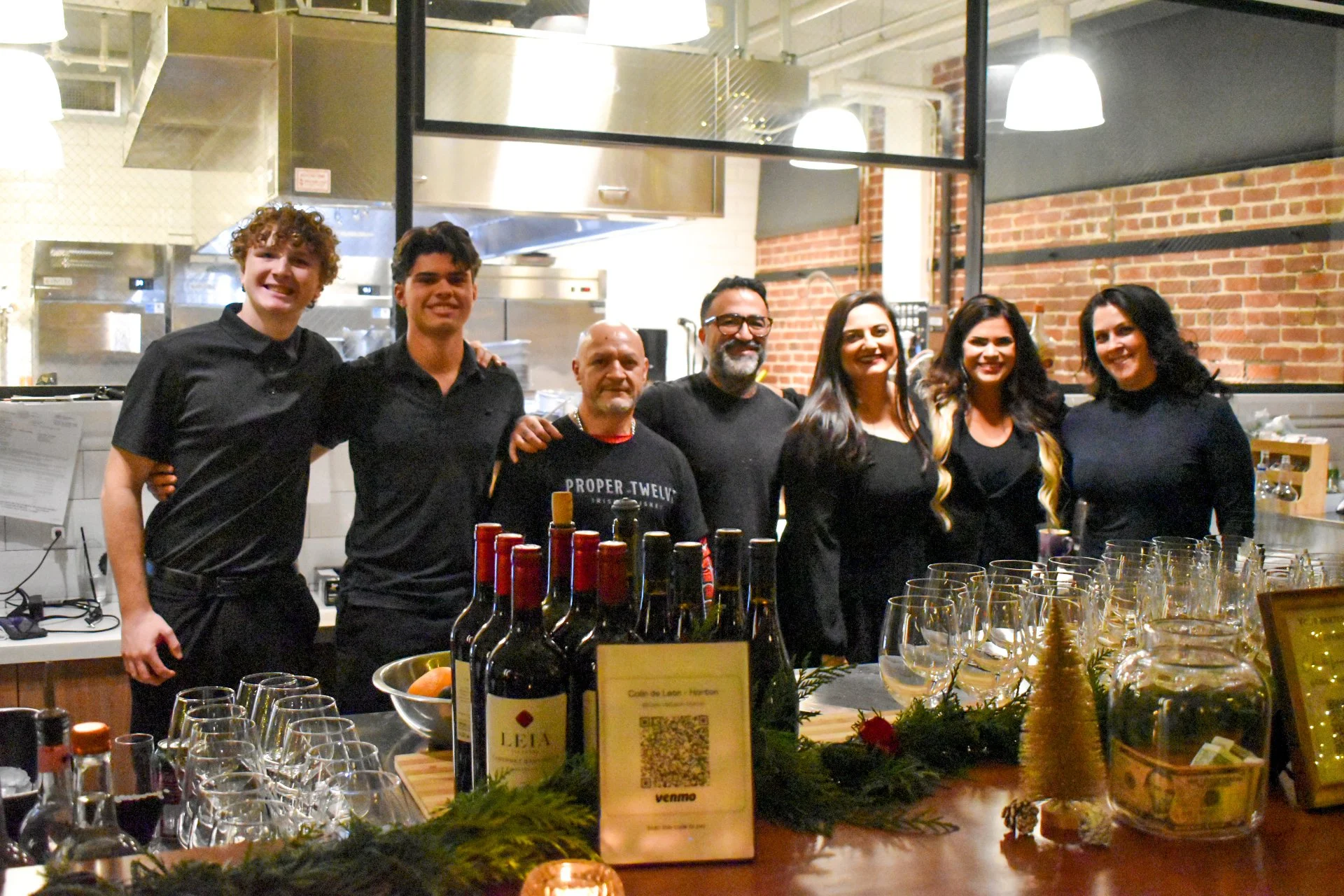 Group of seven people standing behind a bar counter, smiling, with bottles of wine and glasses in front, in a restaurant or bar setting with brick wall and industrial lighting.