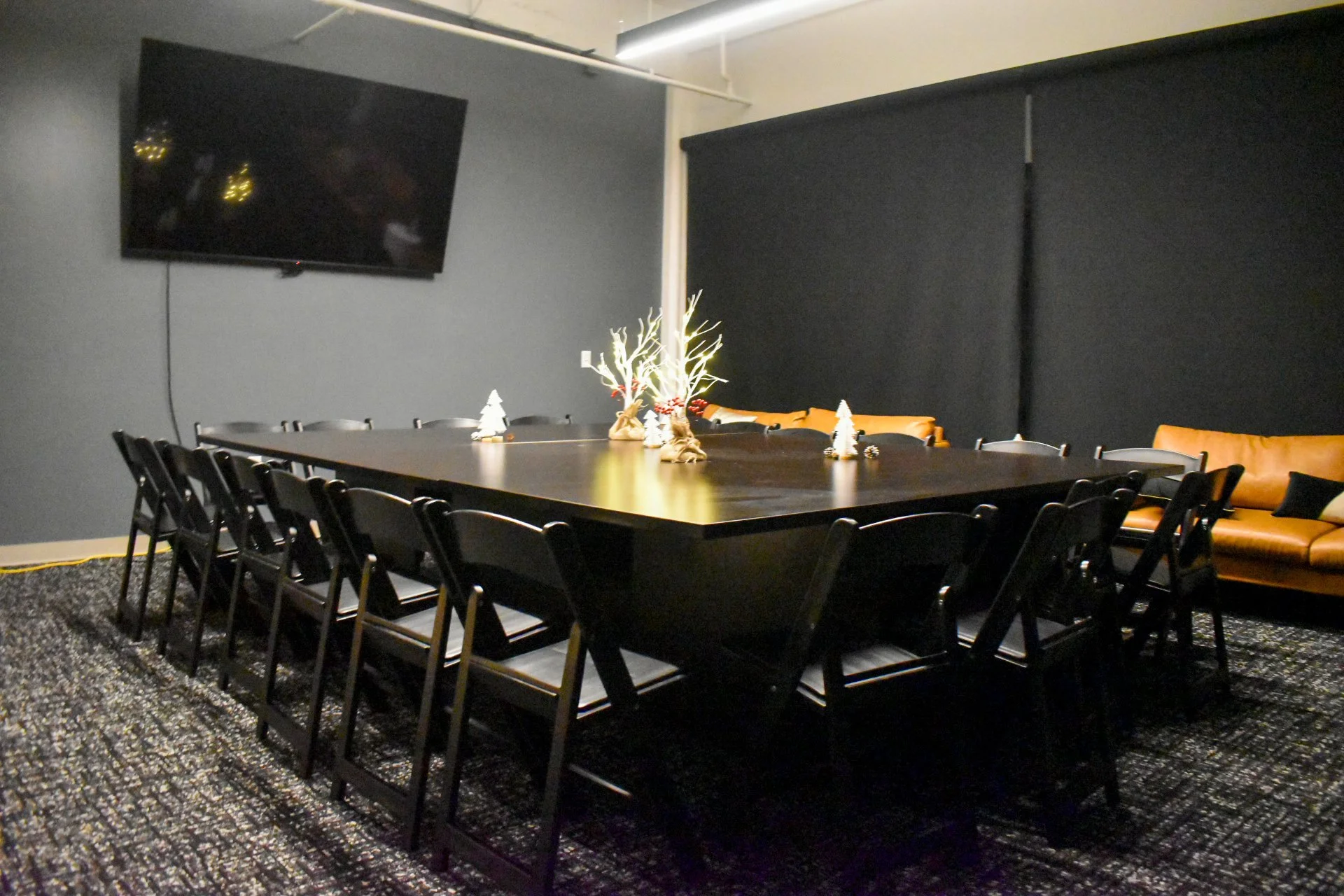 A conference room with black chairs around a large dark table, decorated with Christmas and winter-themed centerpieces, featuring small white Christmas trees and a white branched centerpiece, with a brown sofa against a dark wall and a wall-mounted TV.