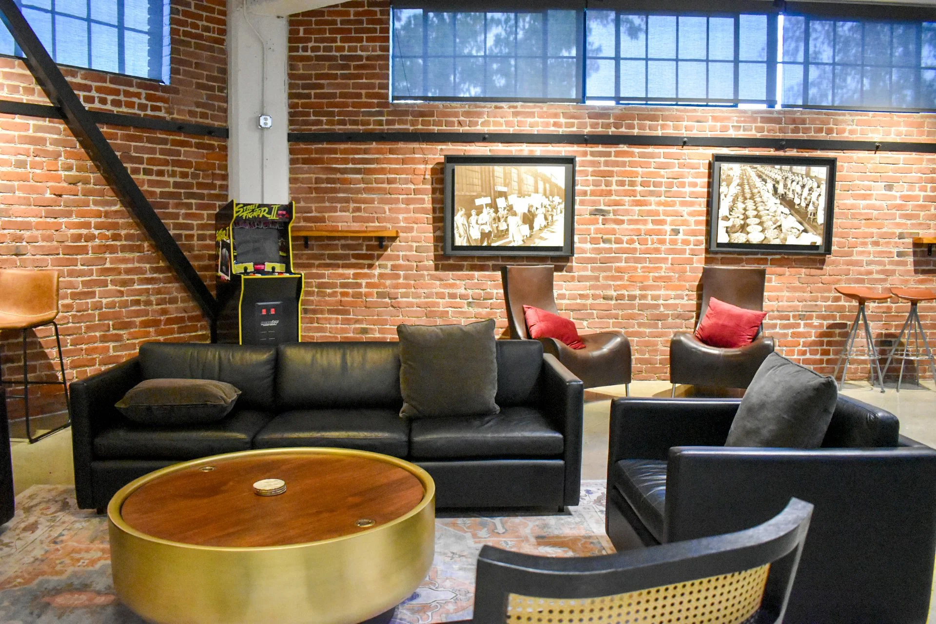 A cozy lounge inside a brick-walled space, featuring black leather sofas and armchairs with cushions, a round brass coffee table, framed vintage photos on the wall, and an arcade Street Fighter II game machine.
