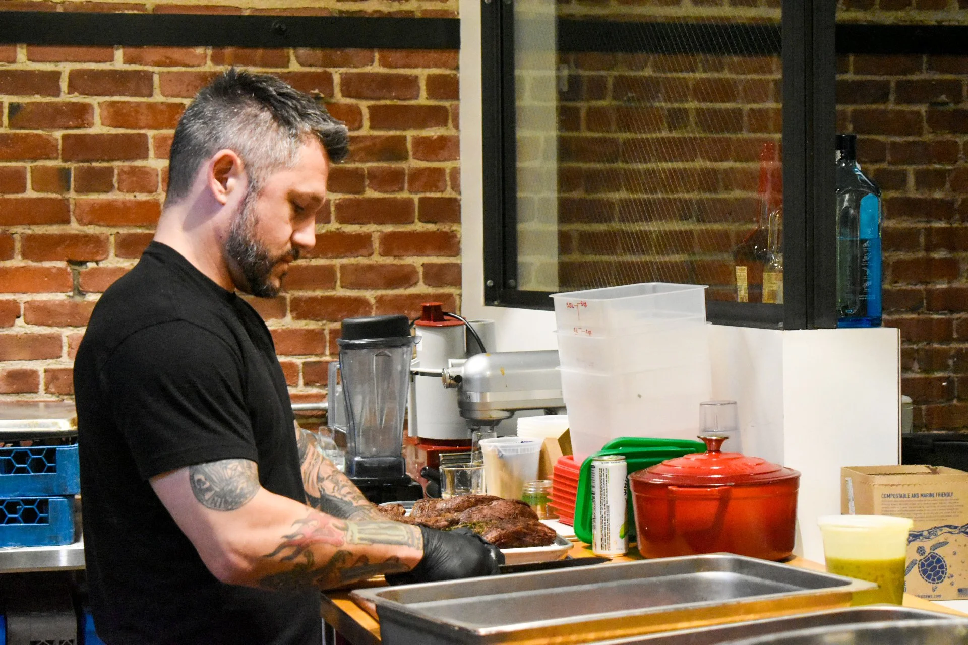 Man with tattoos and black shirt preparing cooked meat in a kitchen with brick wall background.