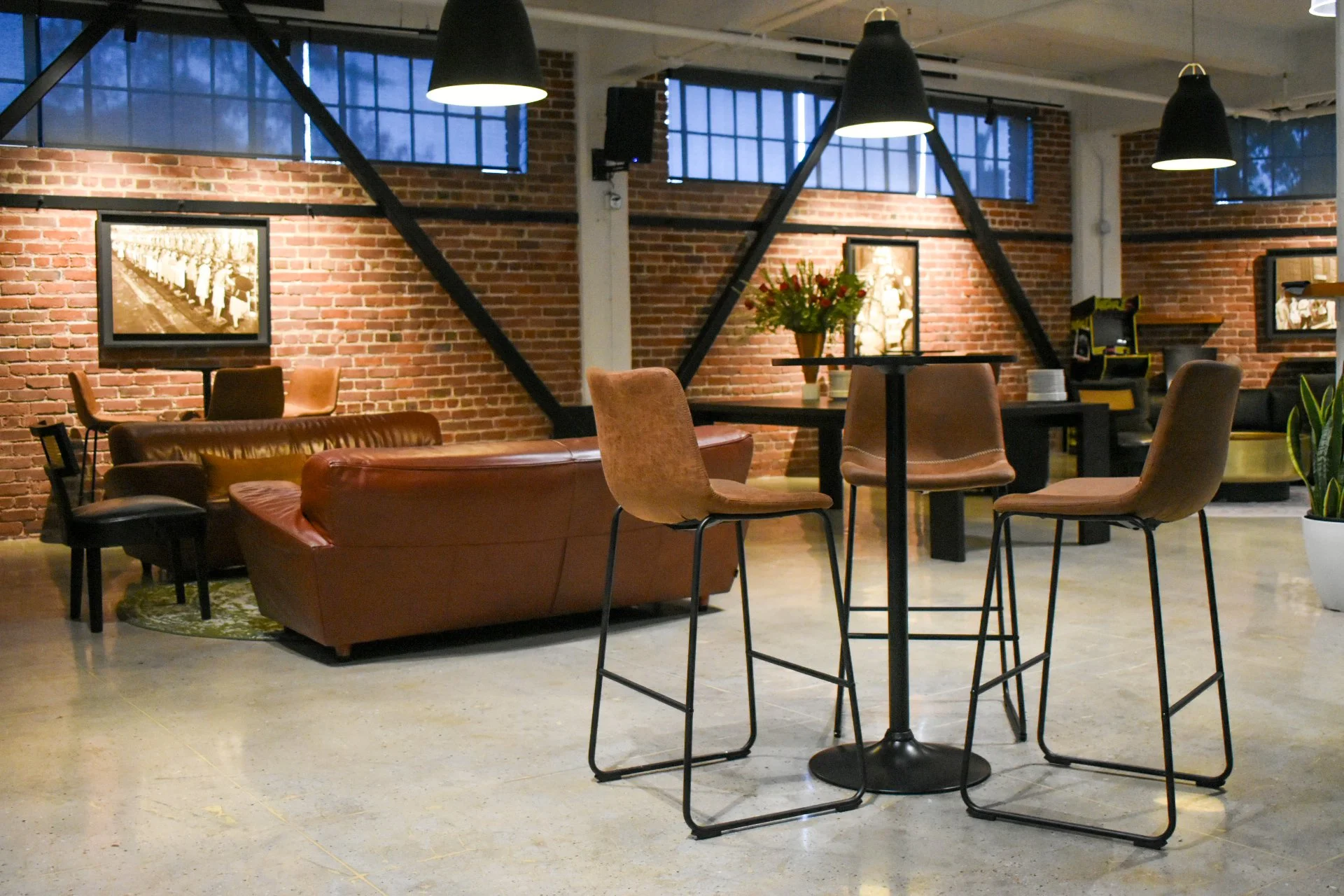 Modern cafe or lounge with brick walls, black pendant lights, leather sofas, and high chairs around a small table, with framed black and white photos on the walls and a potted plant.