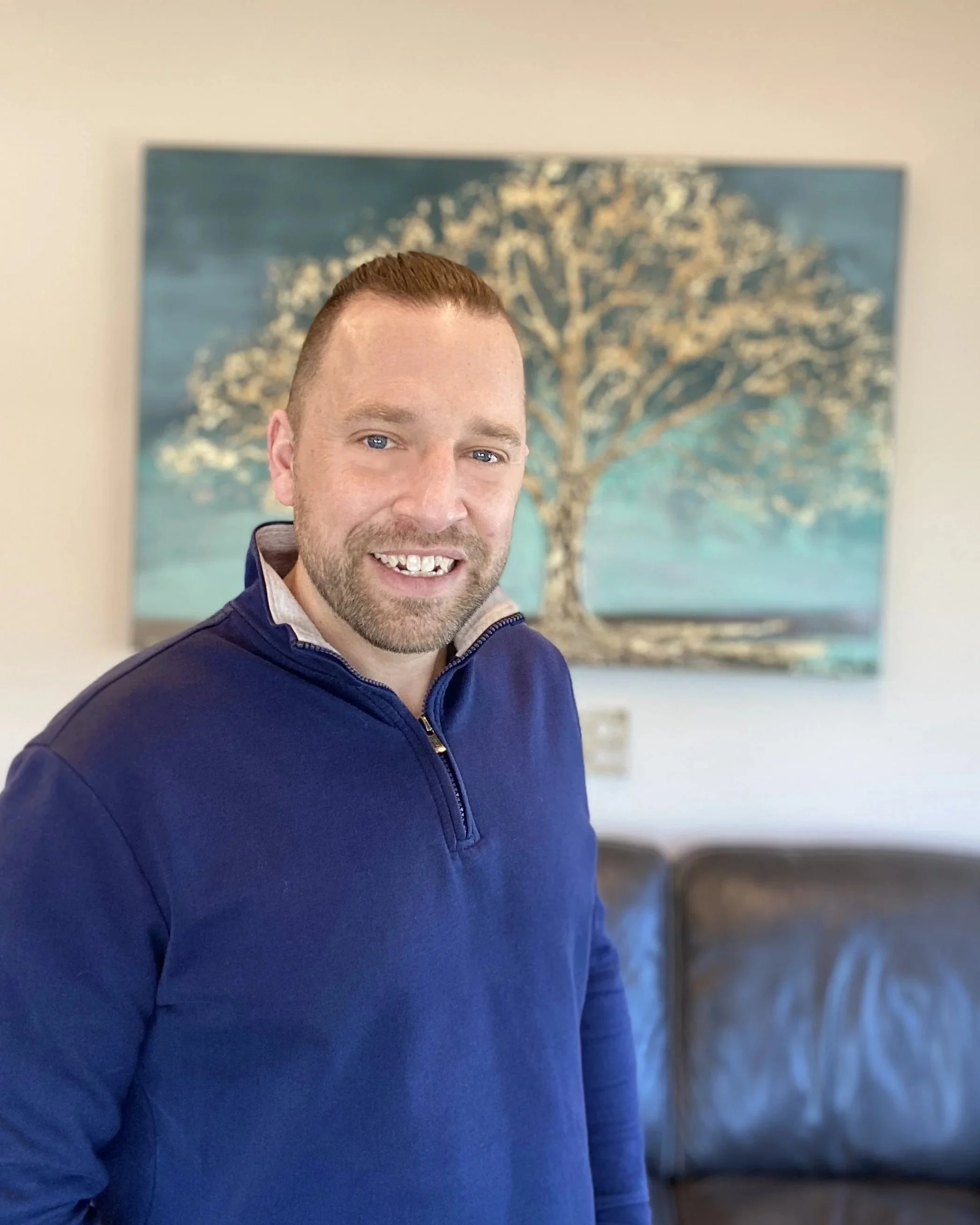 Smiling man in a navy quarter-zip sweater standing in front of a colorful tree painting