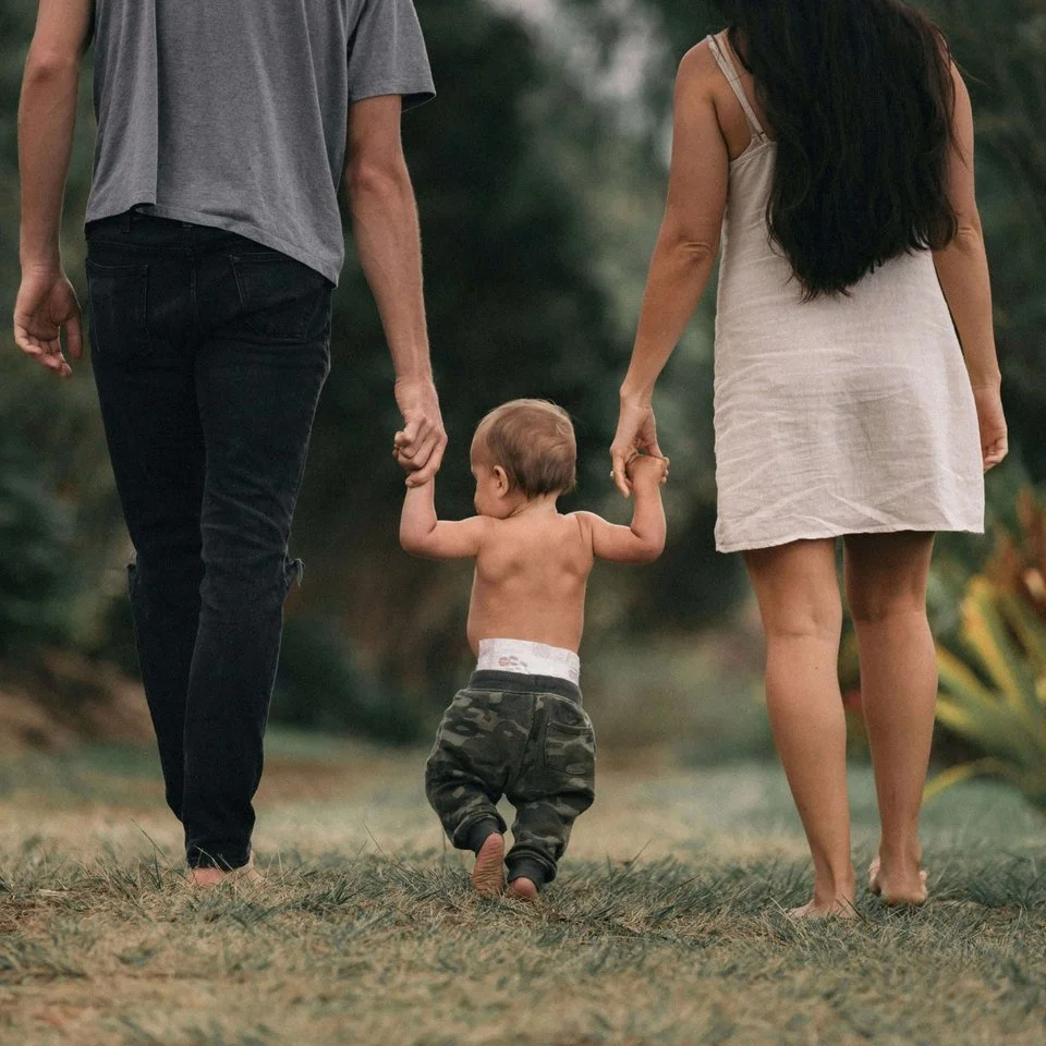 Two adults holding a young child’s hands while walking outdoors.