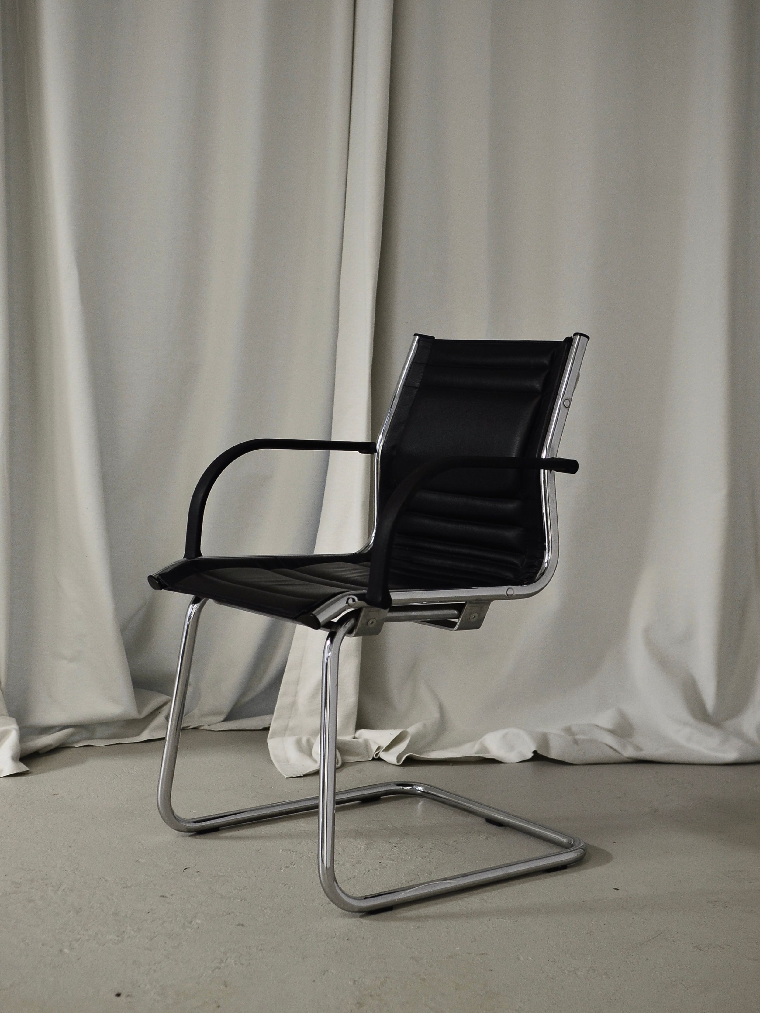 Black leather office chair with metal frame and armrests, positioned in front of white curtains on a neutral carpeted floor.