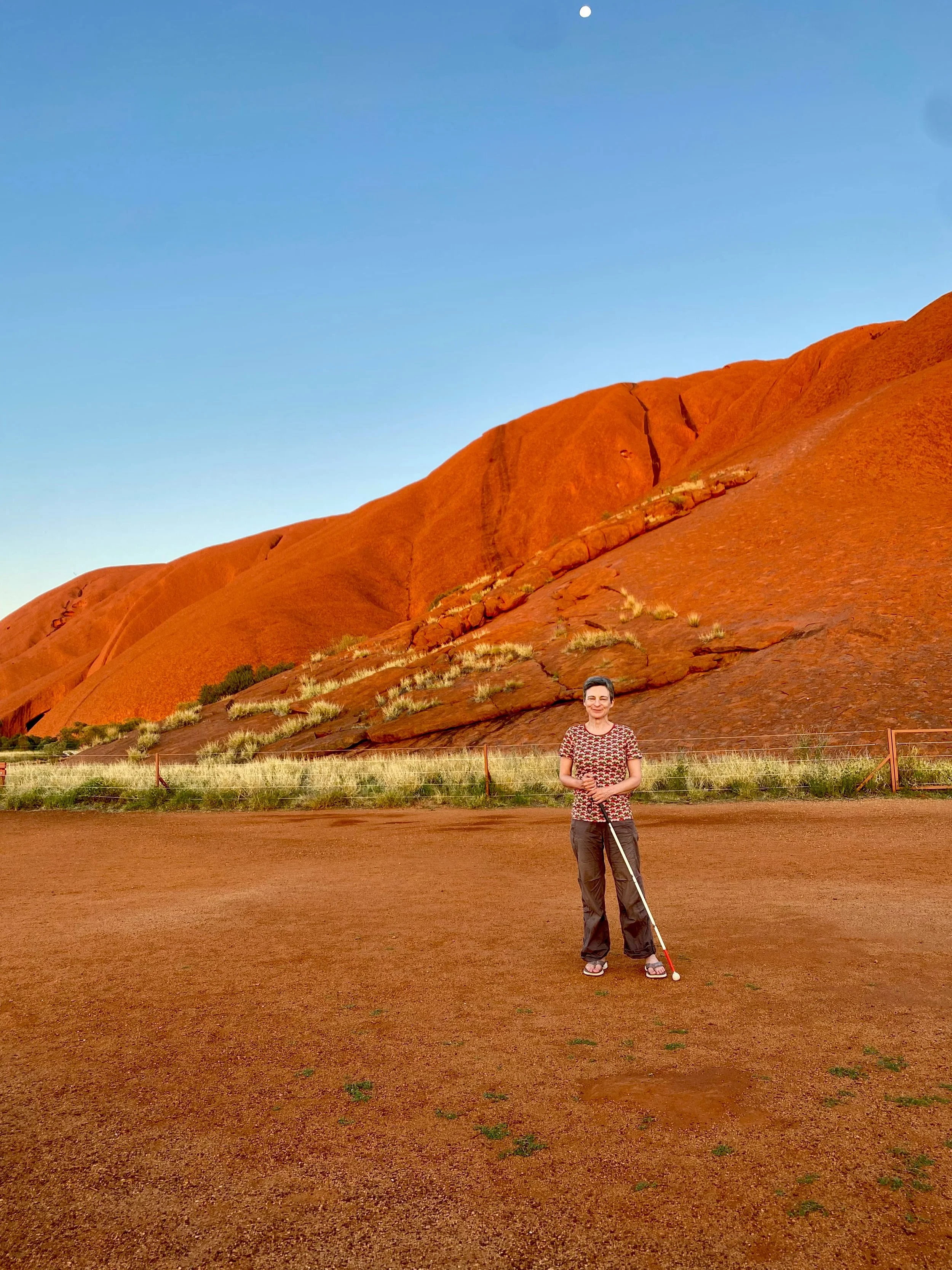 Llewelyn smiles, holding her cane, standing in front of the natural rich red/orange colour of Uluru behind her.