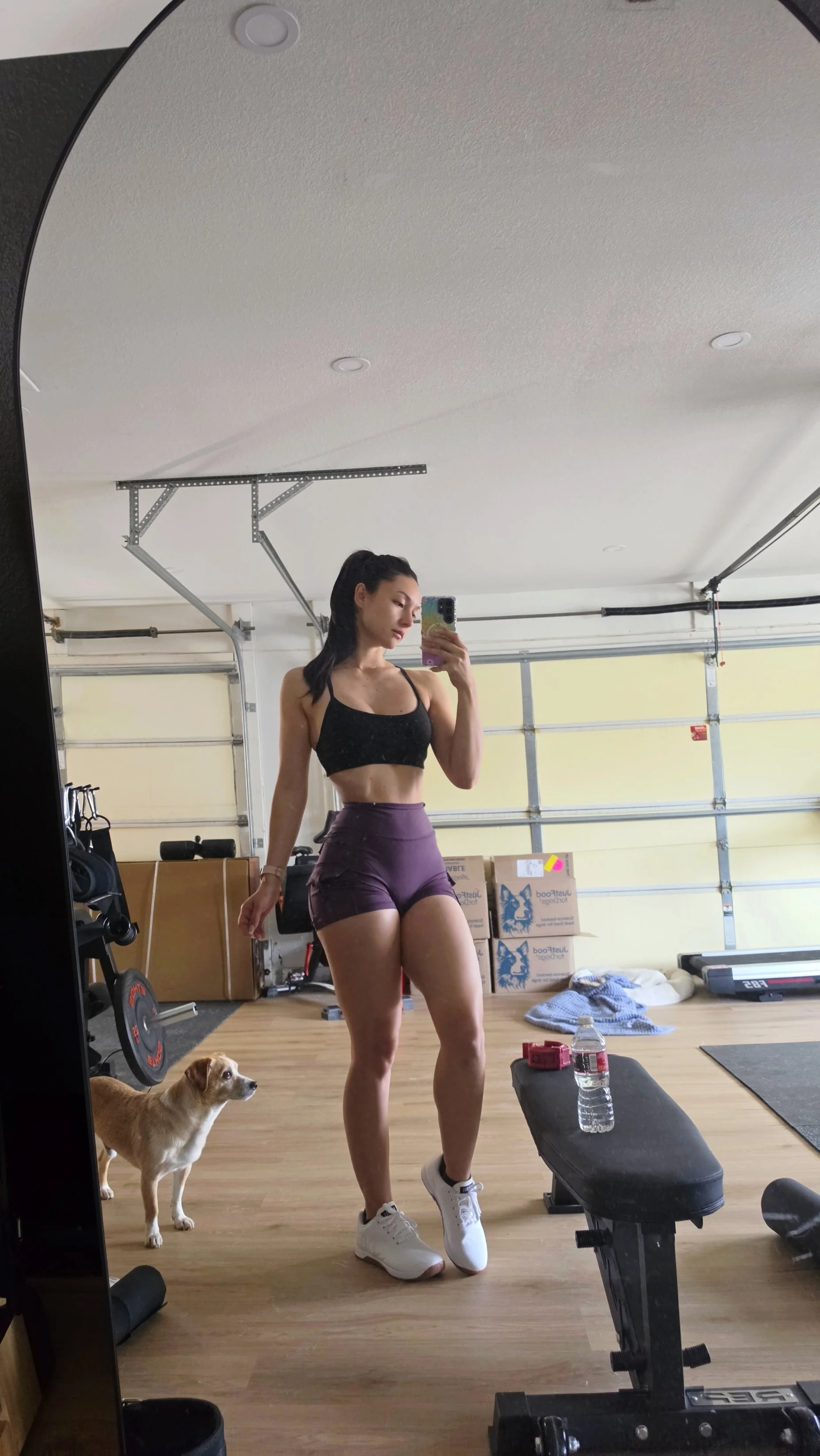 Cailey in her garage home gym with her dog watching from beside the rack