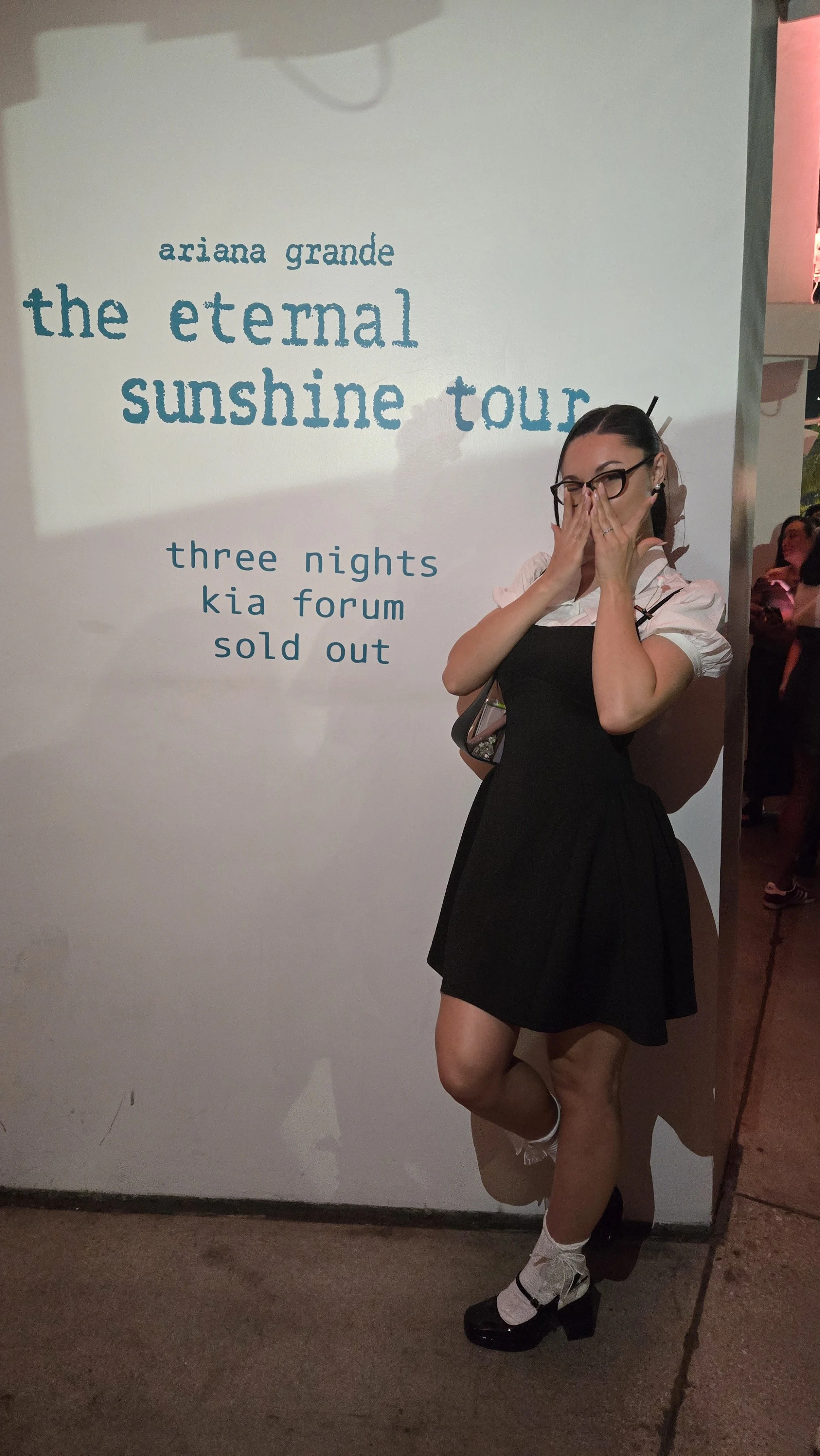 Standing in front of the Ariana Grande Eternal Sunshine Tour mural in a black dress over a white puff sleeve top and cat eye glasses