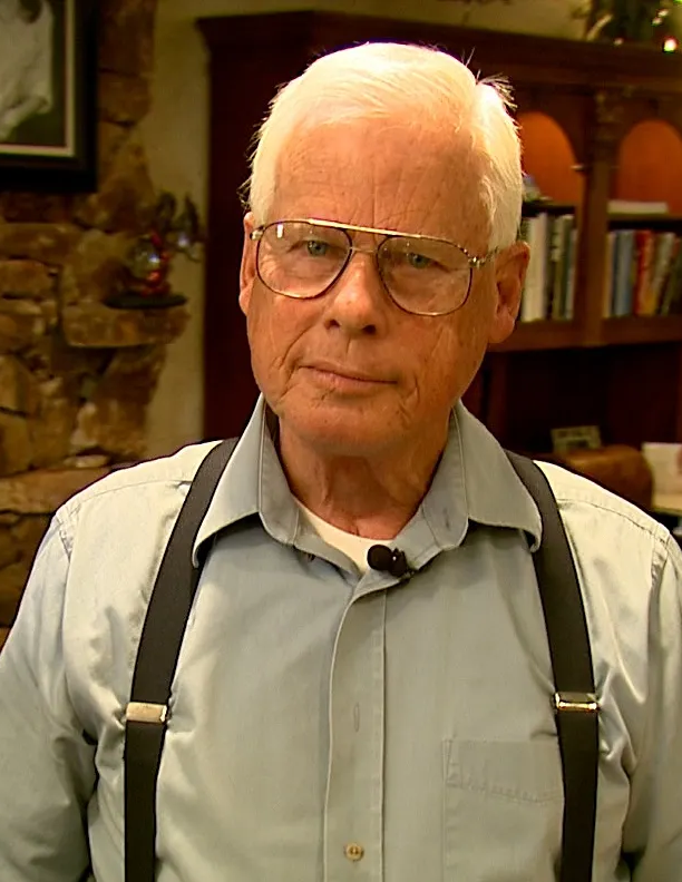 Elderly man wearing glasses and a light blue shirt with suspenders, standing indoors, with bookshelves and stone wall in the background.