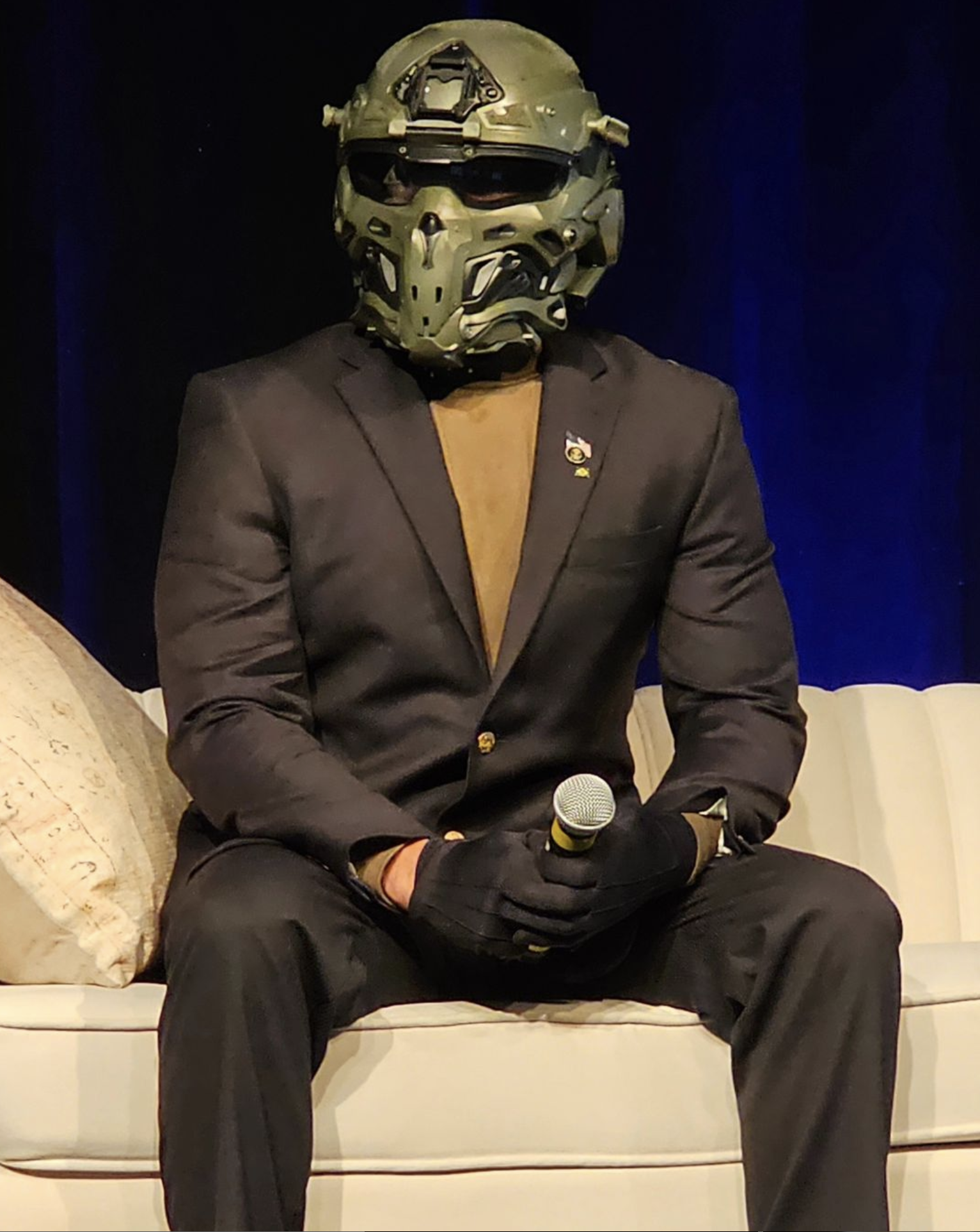 Person in a suit wearing a green sci-fi helmet and black gloves, holding a microphone, seated on a beige sofa.