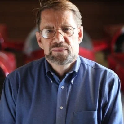 Man wearing glasses and a blue button-up shirt standing indoors with a blurred background.