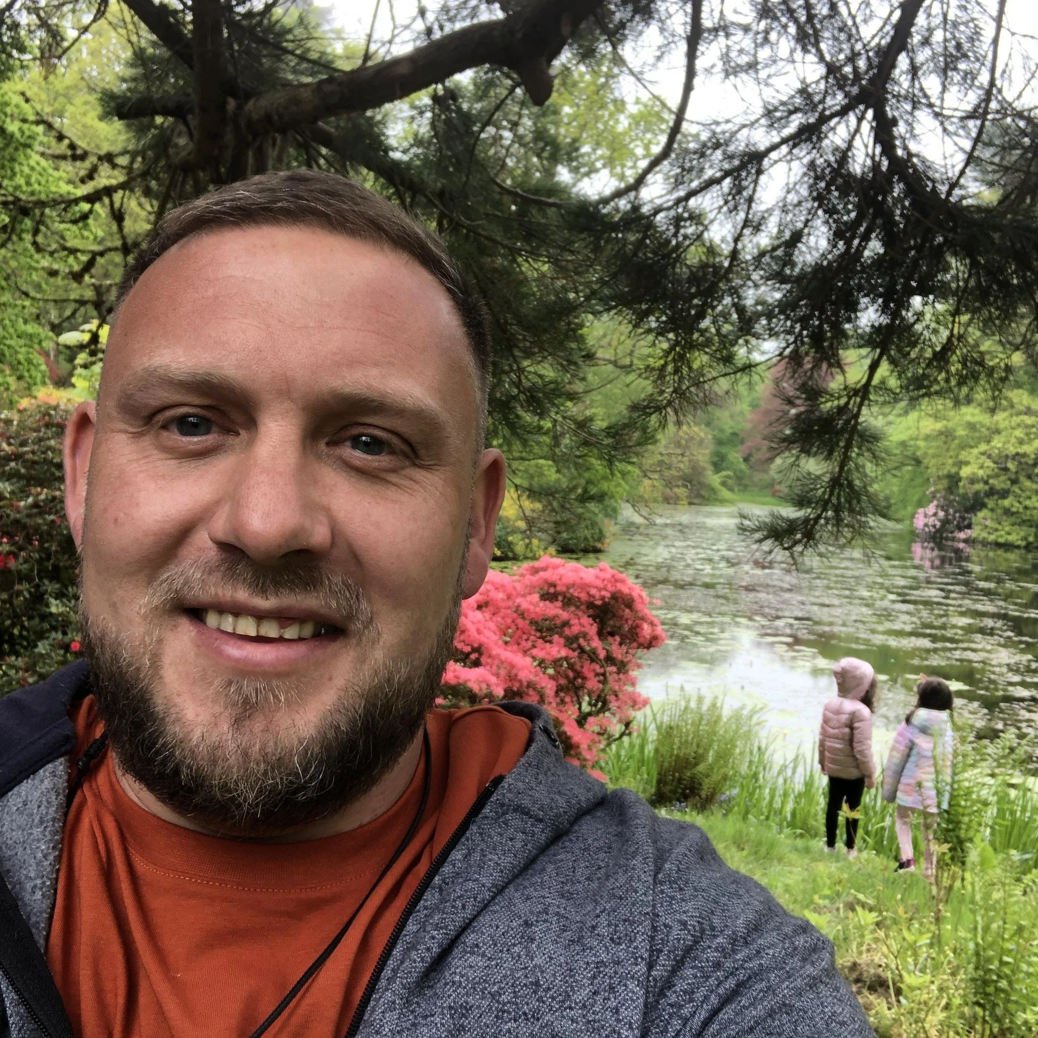 A man taking a selfie in a lush garden with a pond and two children in the background. The garden is vibrant with greenery and flowering plants, including pink azaleas. The children are standing near the water, both wearing hooded jackets.