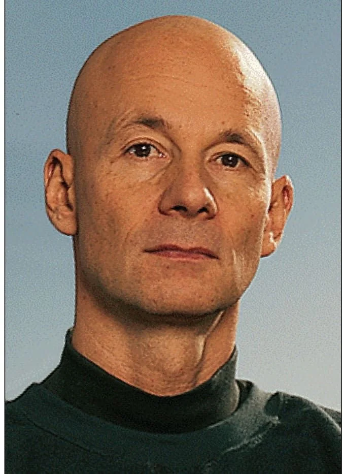 Close-up portrait of a bald man with a neutral expression, wearing a dark turtleneck, against a light blue background.