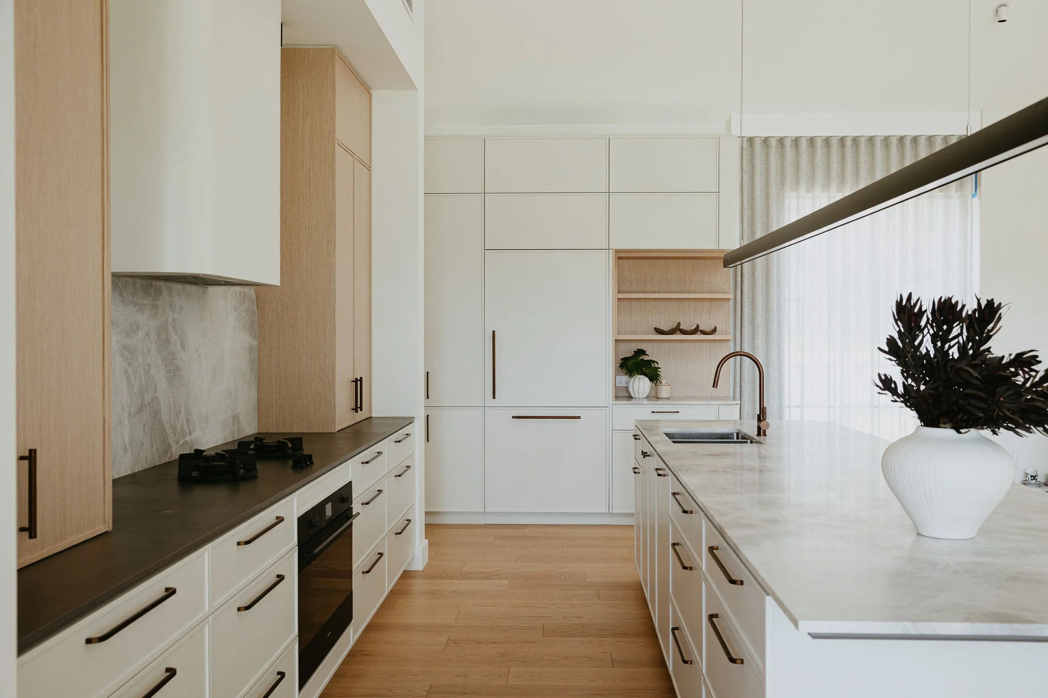 Light-filled luxury kitchen featuring integrated appliances, a stone island benchtop, and warm wood finishes, blending contemporary design with everyday functionality.