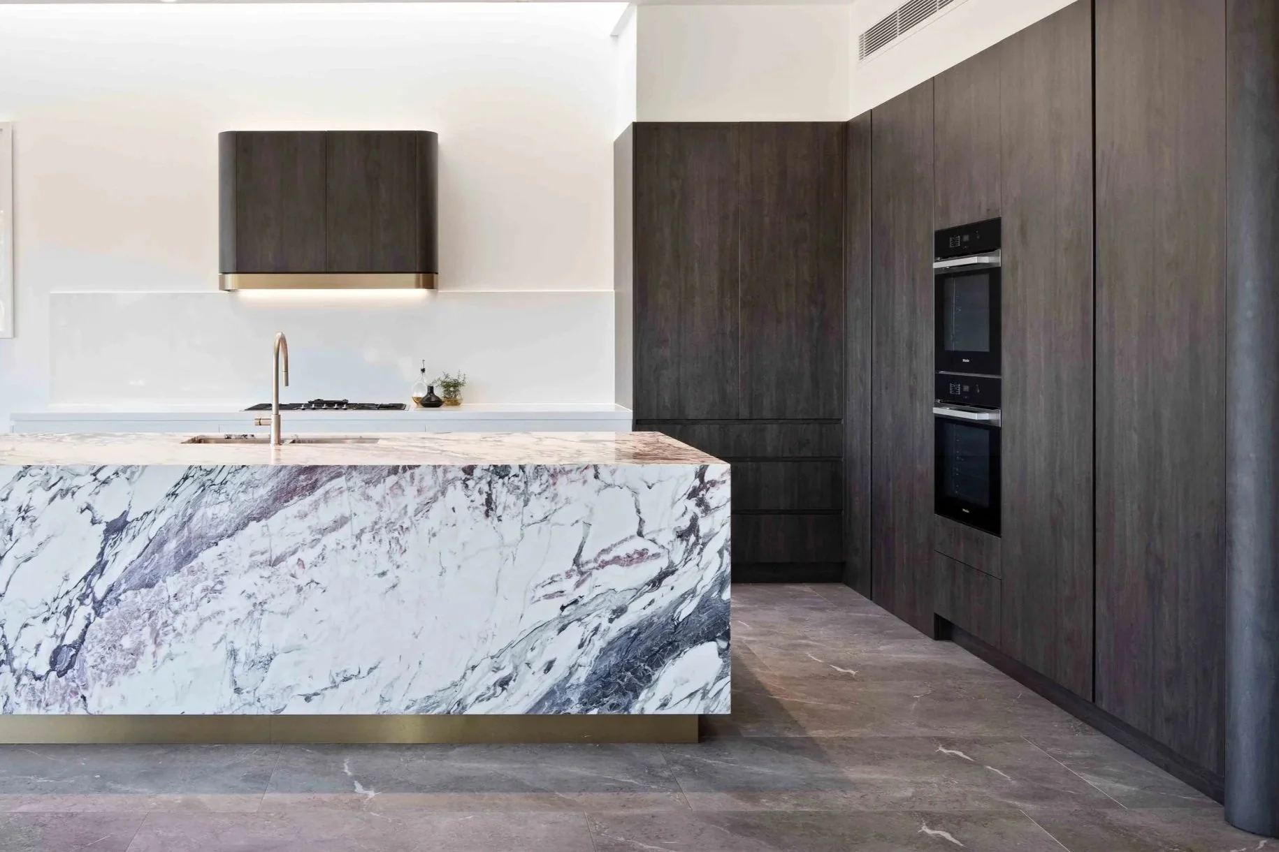 Custom chocolate veneer rangehood with gold edge detailing - dark wood veneer joinery cabinetry with intergrated fridge and freezer. Featuring oversized natural stone black island bench with gold brass kickboard and gold sink