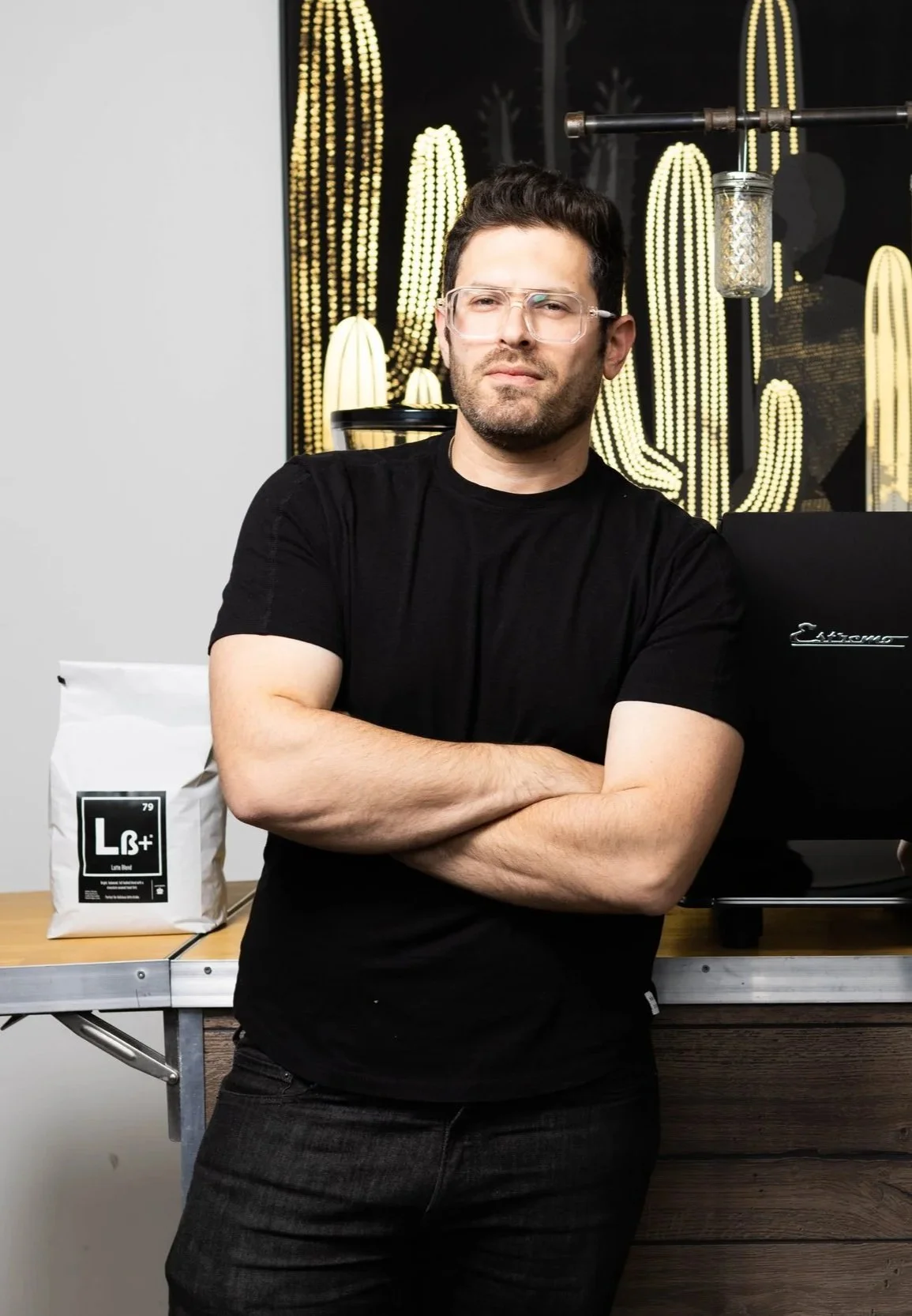 Our company's founder standing in front of a mobile coffee bar setup, featuring coffee equipment and a black espresso machine, with wall-mounted shelves displaying various coffee-making items; both men are wearing black t-shirts.