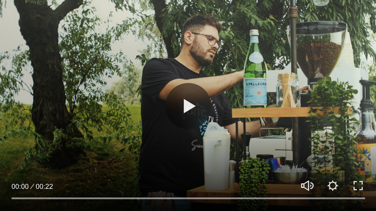 A man making coffee outdoors at a mobile coffee stand with trees in the background.