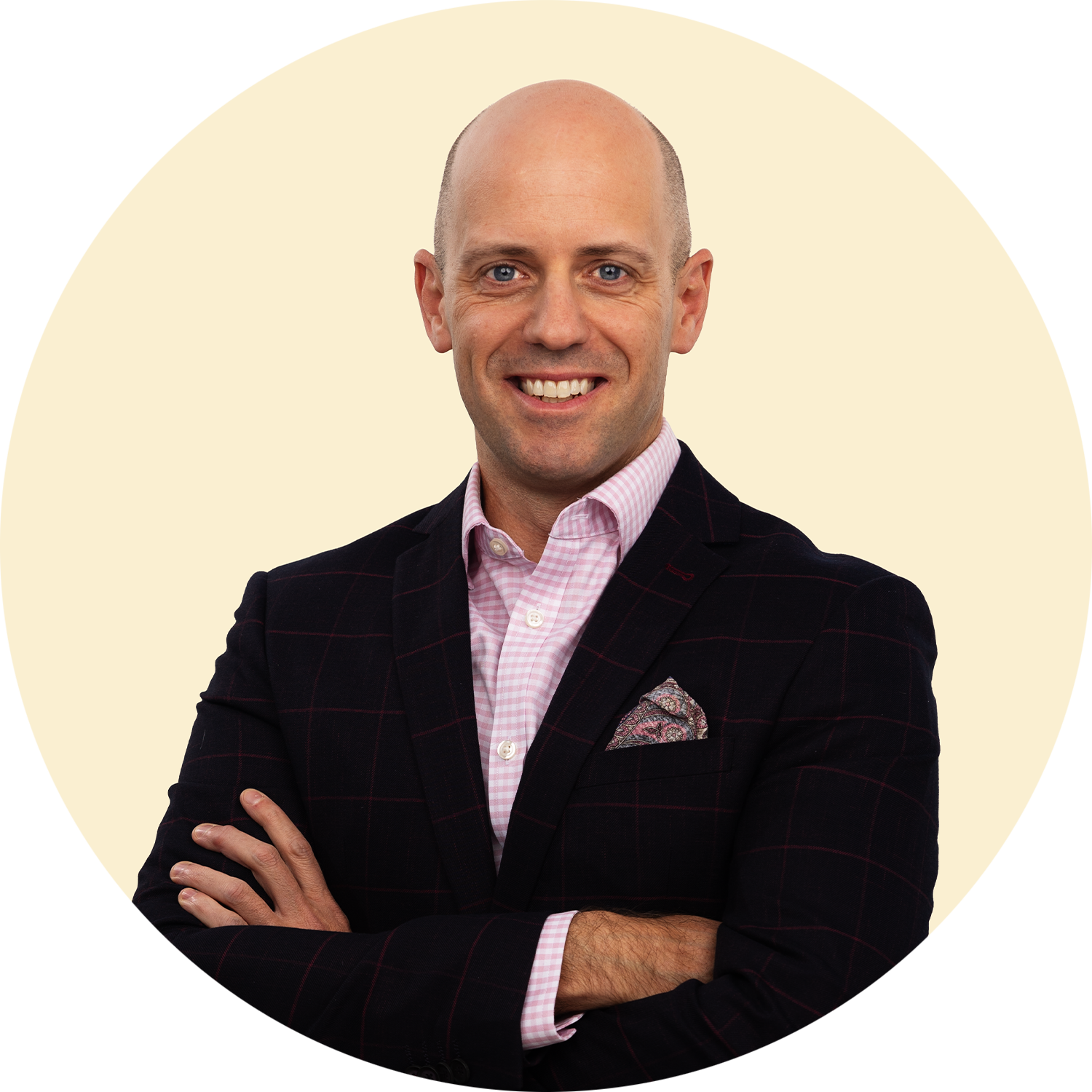 Headshot of a smiling man with a bald head, wearing a dark plaid blazer, pink gingham shirt, and a pocket square, standing with arms crossed against a light background.