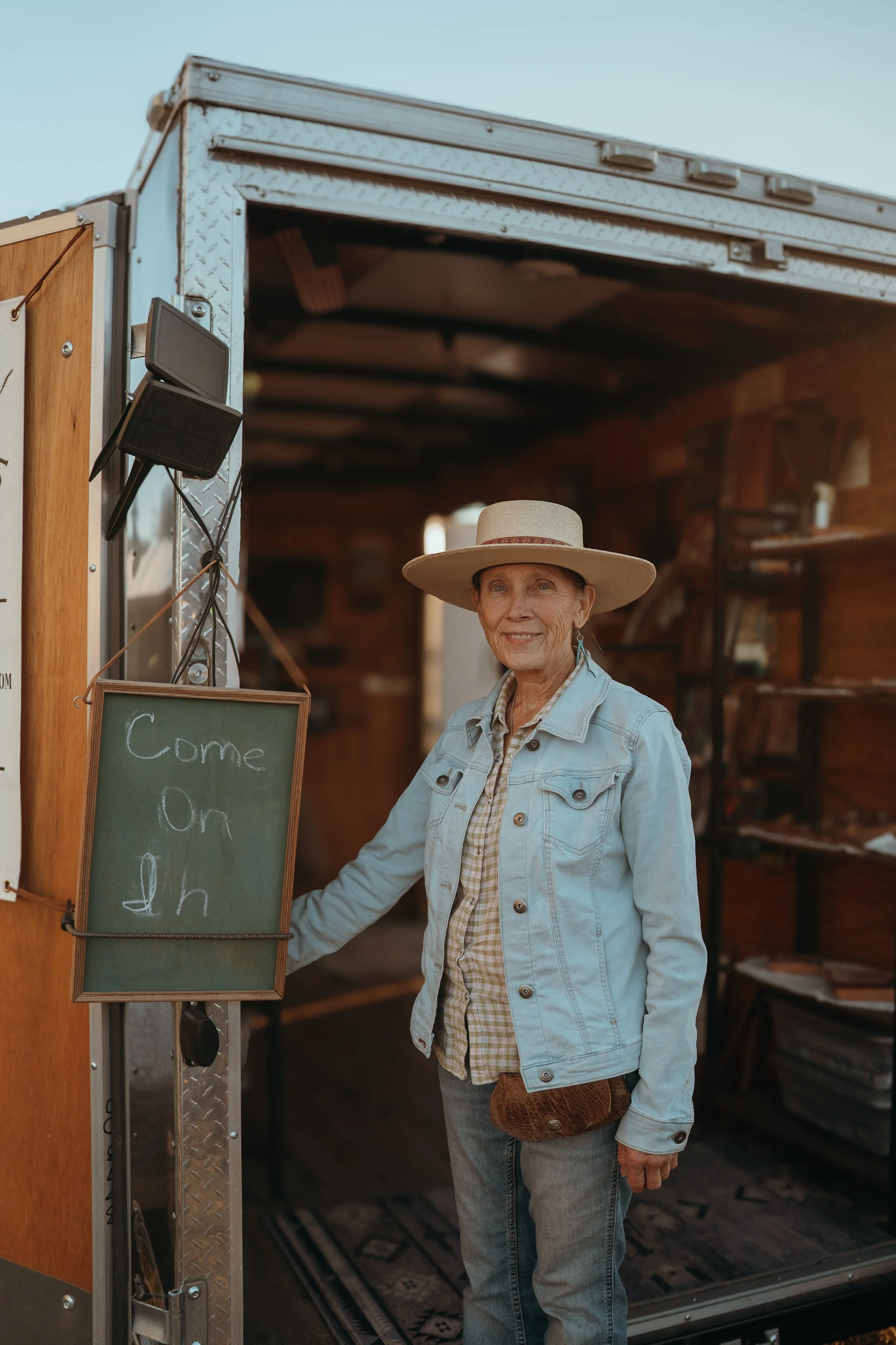 A woman in a white shirt and blue jeans holding a corn dog in front of a food stall with cotton candy and lollipops at a fair or carnival.