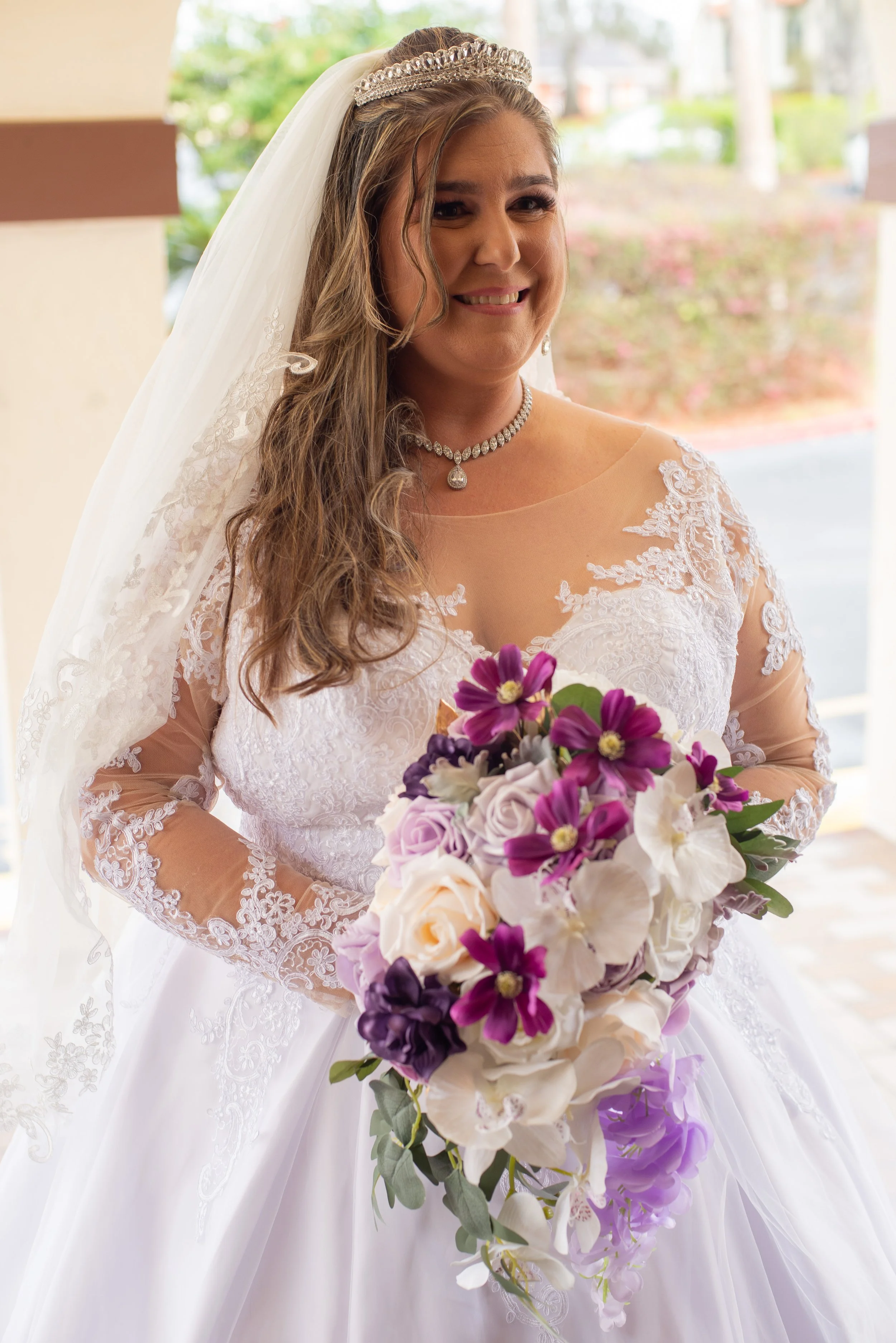 A bride in a white wedding gown with lace detailing, wearing a tiara and a pearl necklace, holding a large bouquet of purple, white, and cream flowers, standing indoors near an open entrance with greenery outside.