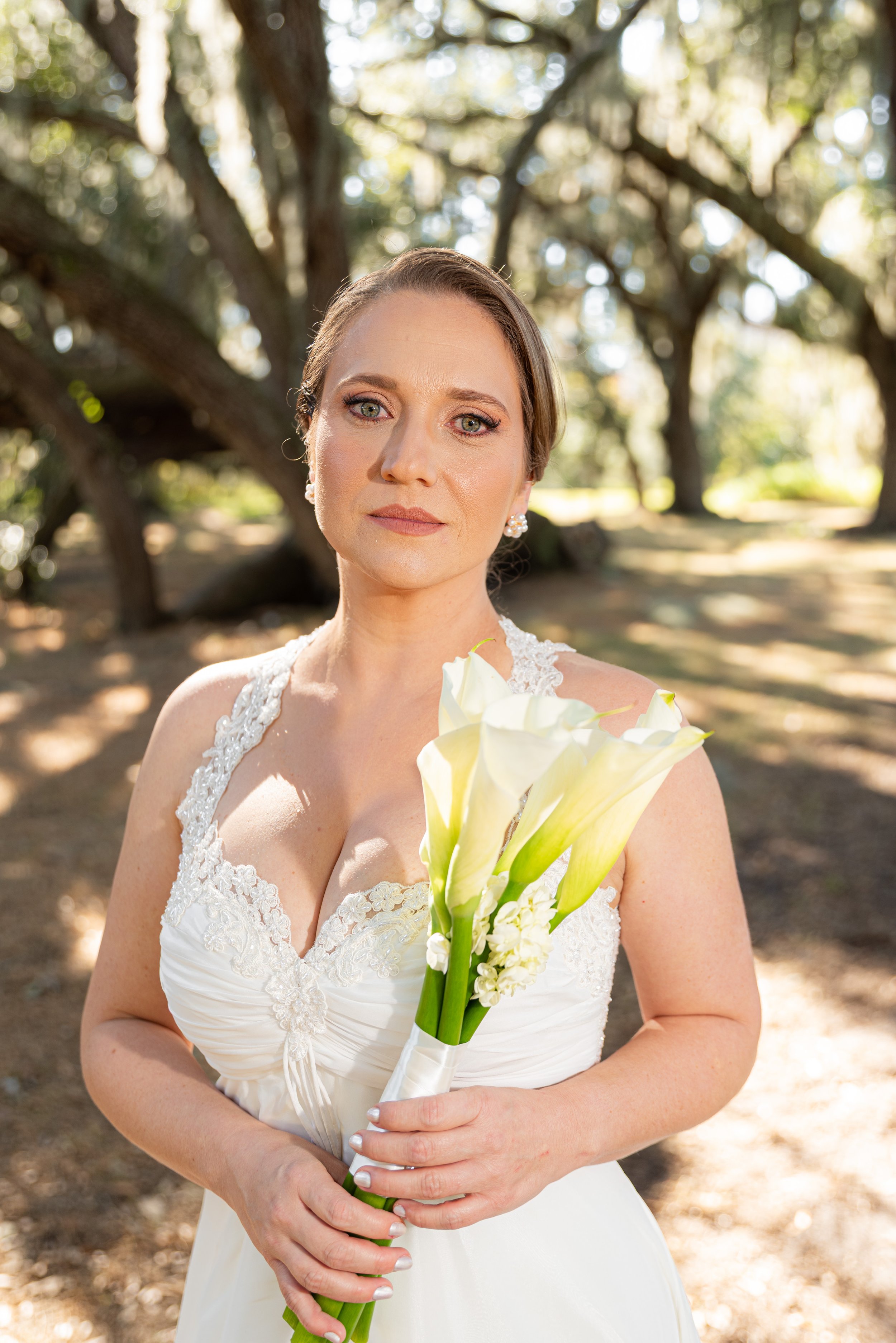 A bride in a white wedding dress holding calla lilies, standing outdoors with trees in the background.