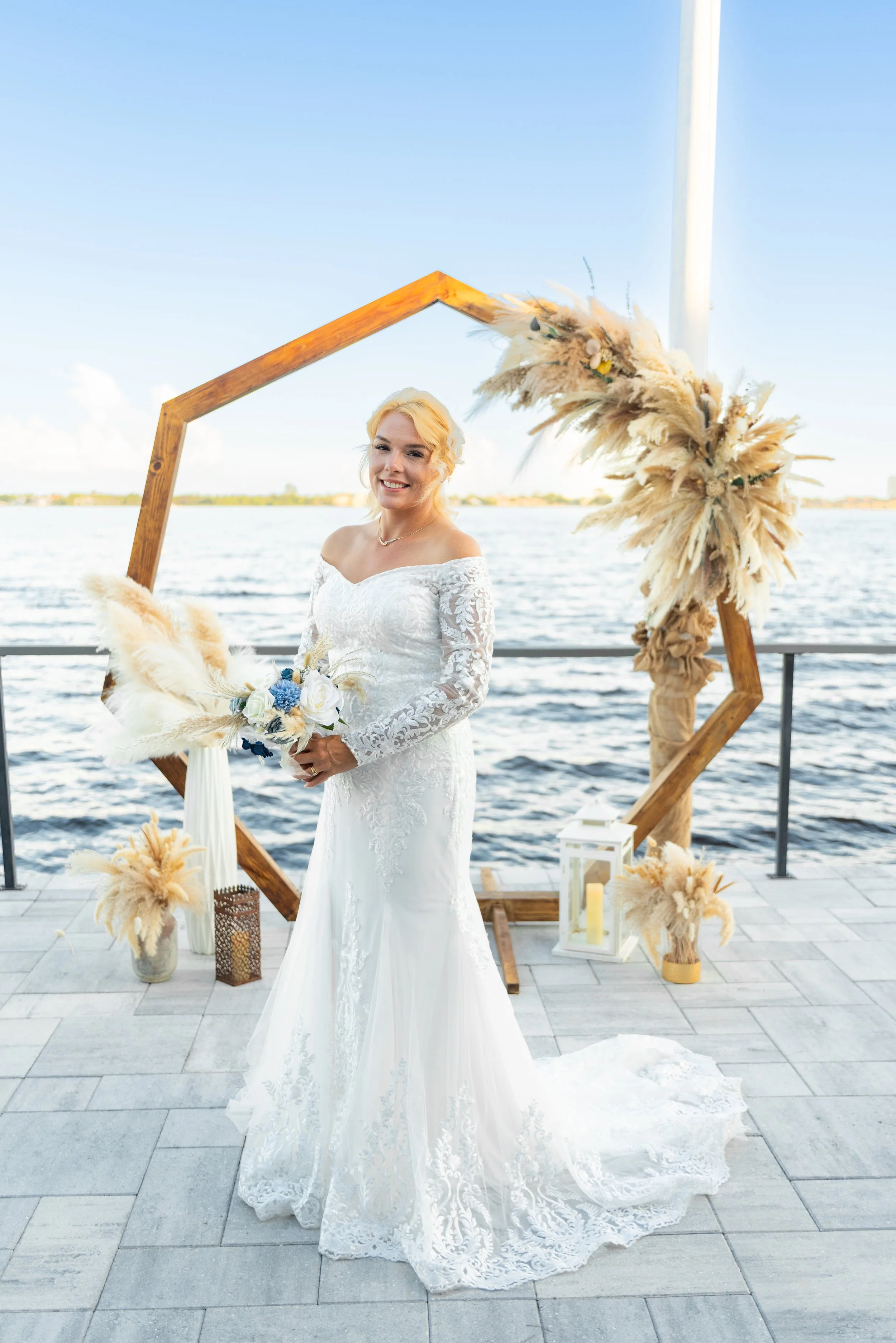 A bride in a white lace wedding dress holding a bouquet, standing outdoors by a body of water with a decorated wooden frame and pampas grass arrangements, near lanterns, under a clear blue sky.