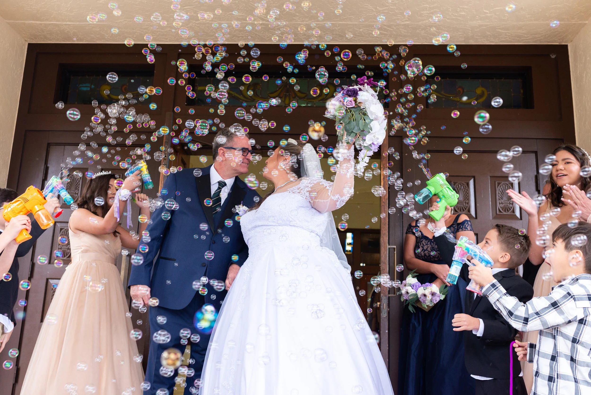 A bride and groom celebrating with bubbles and water guns, surrounded by children and guests outside a church.