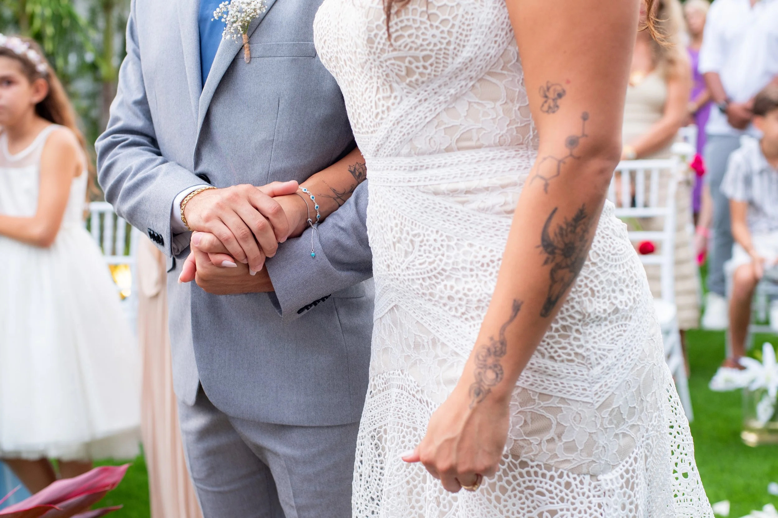A couple holding hands at a wedding ceremony with several guests in the background. The man wears a light gray suit, and the woman wears a white lace dress with tattoos on her arm.